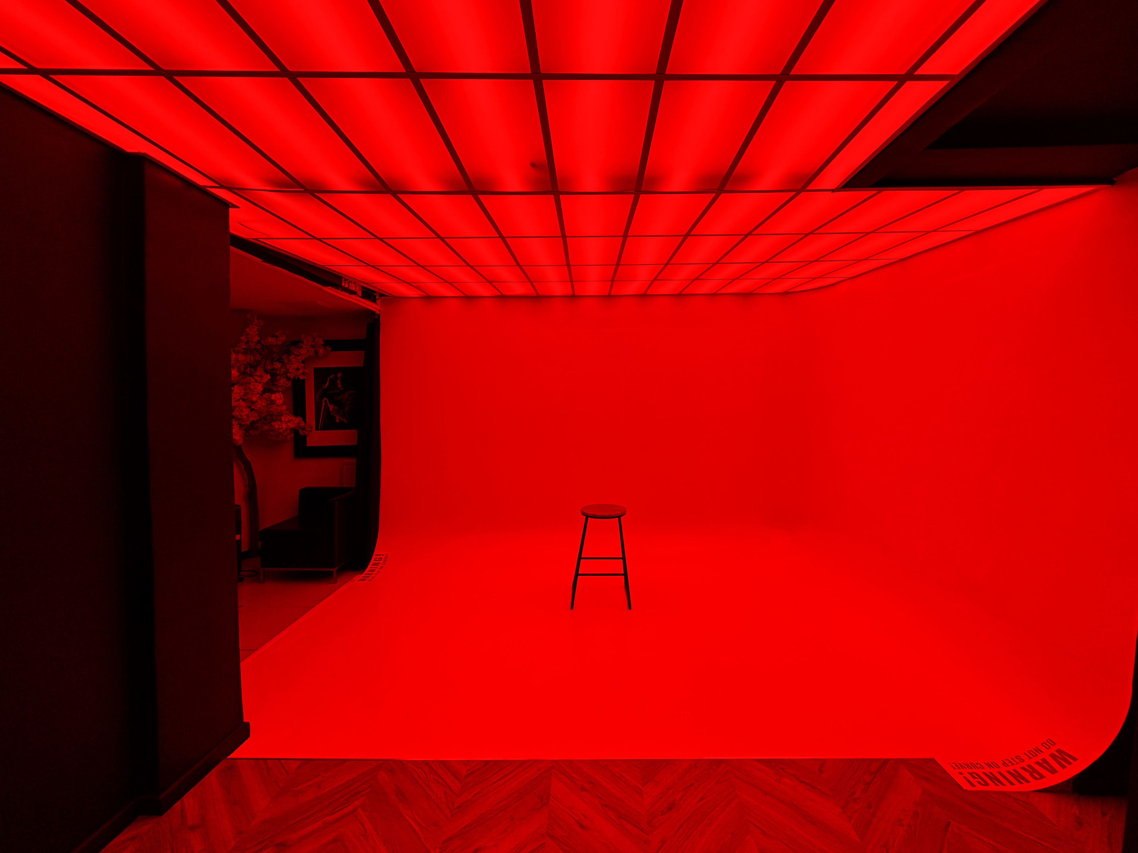 A minimalist photo studio features a red backdrop and a single wooden stool positioned in the center.