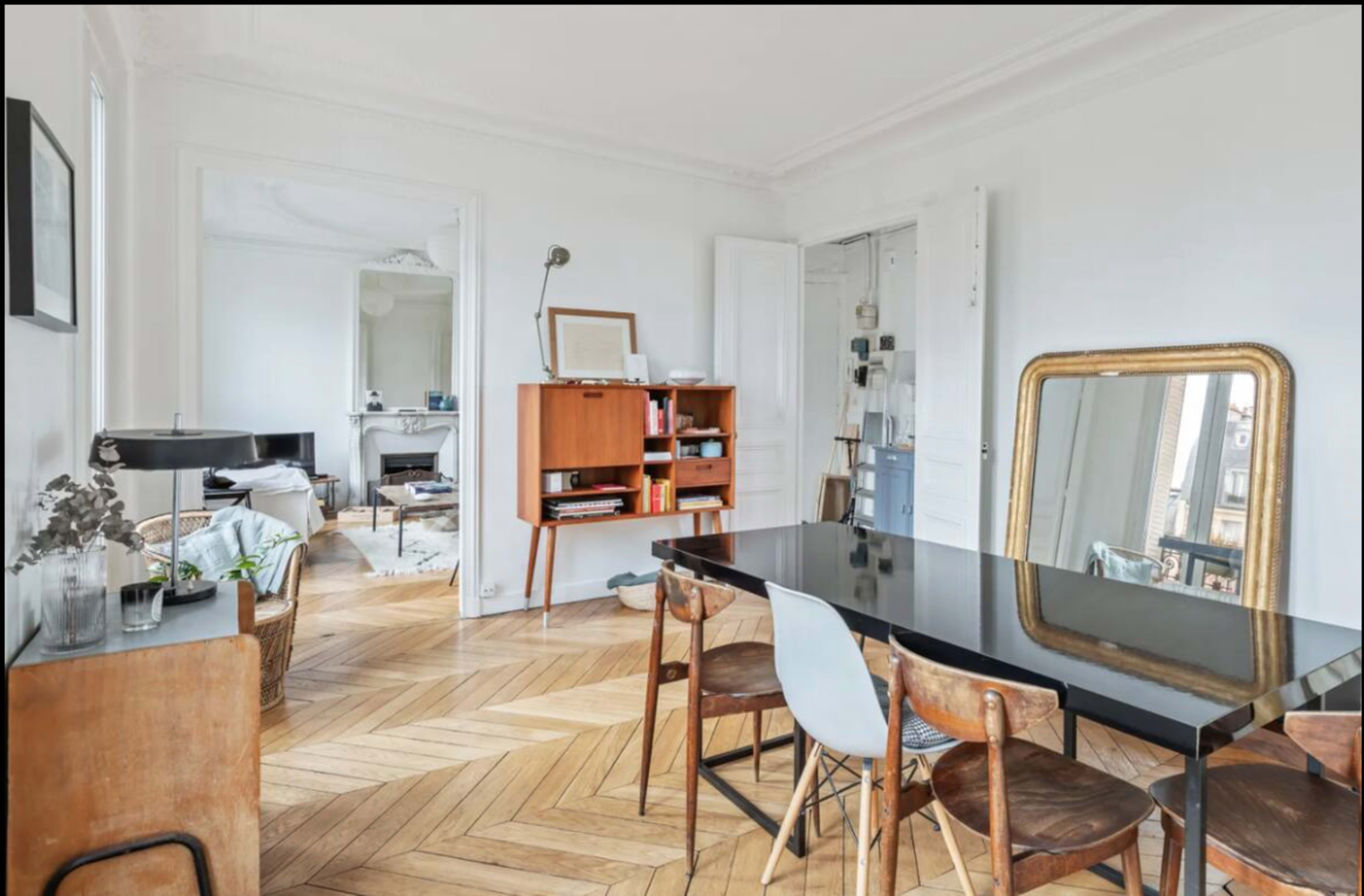 The image shows a modern living space with a black dining table surrounded by wooden chairs, a minimalist bookshelf, and large windows that provide natural light.