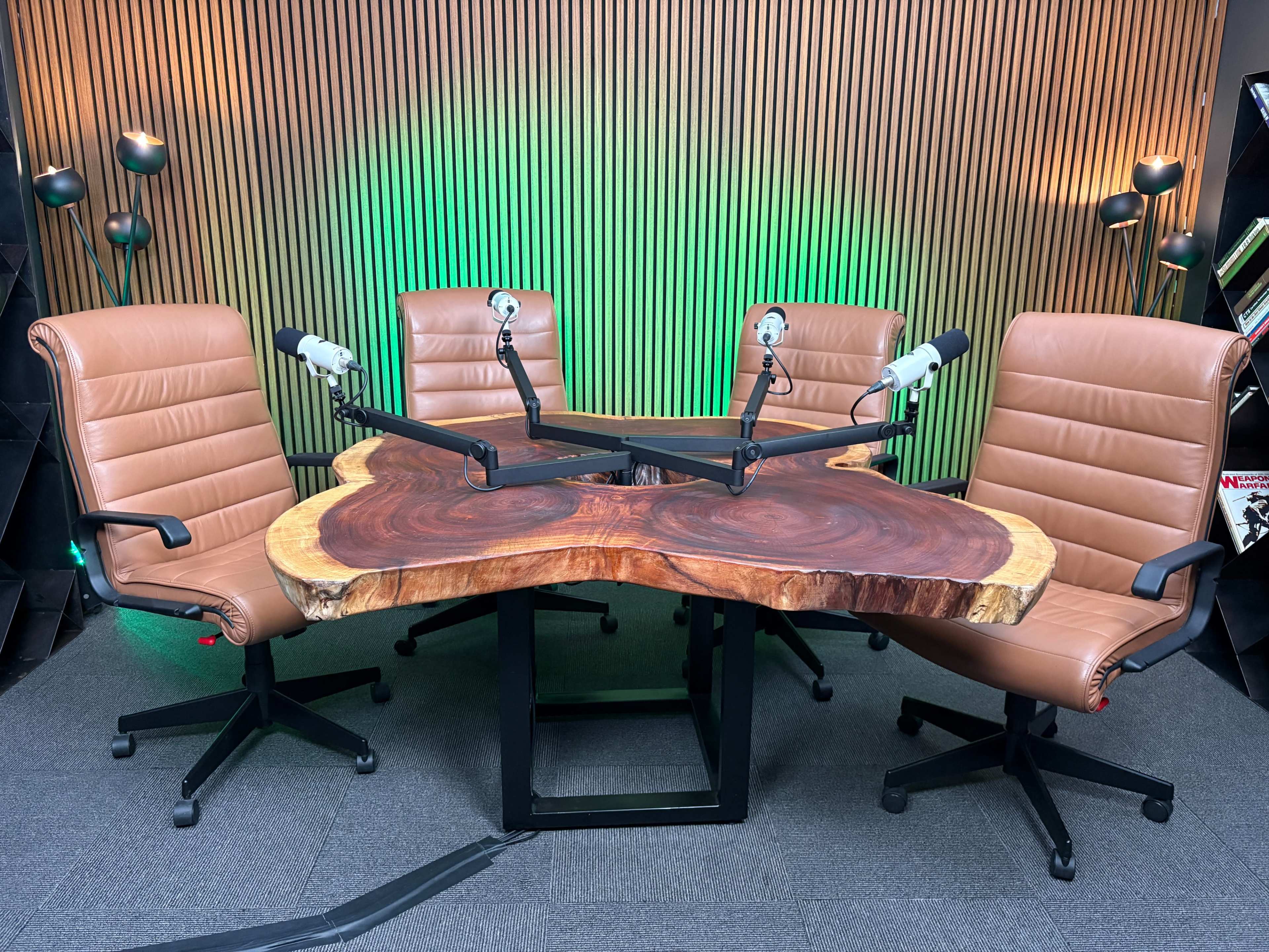 A custom wooden table with a natural edge is surrounded by four brown leather office chairs, each equipped with a microphone, set against a backdrop of wooden paneling and green lights.