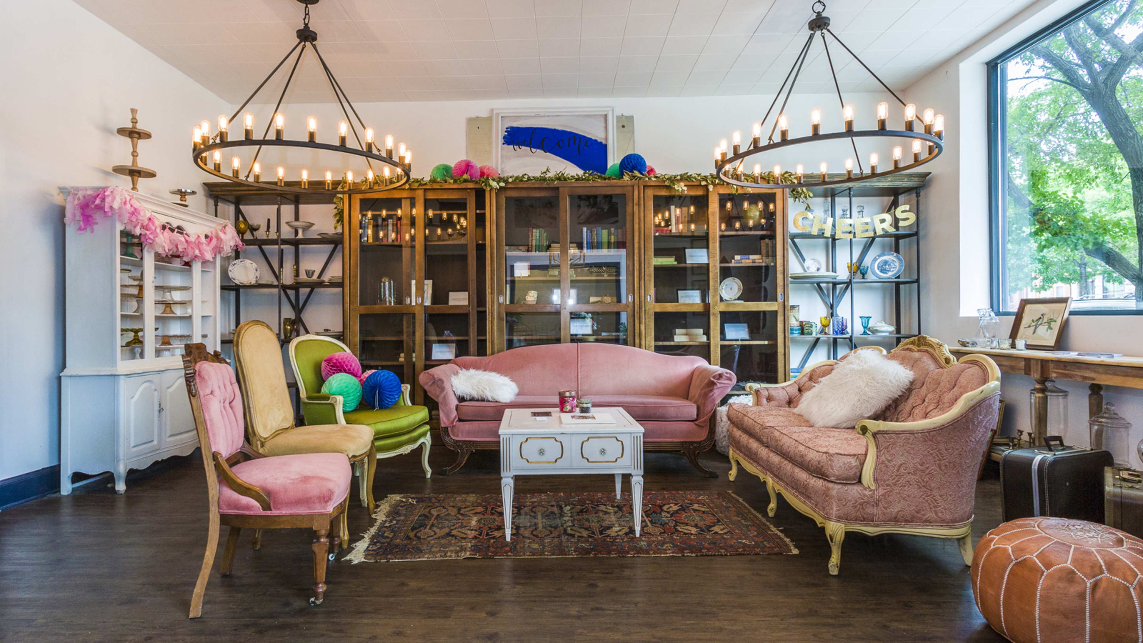 The scene features a stylishly arranged living area with vintage furniture, including a pink sofa, green chair, and a coffee table, illuminated by large chandeliers.