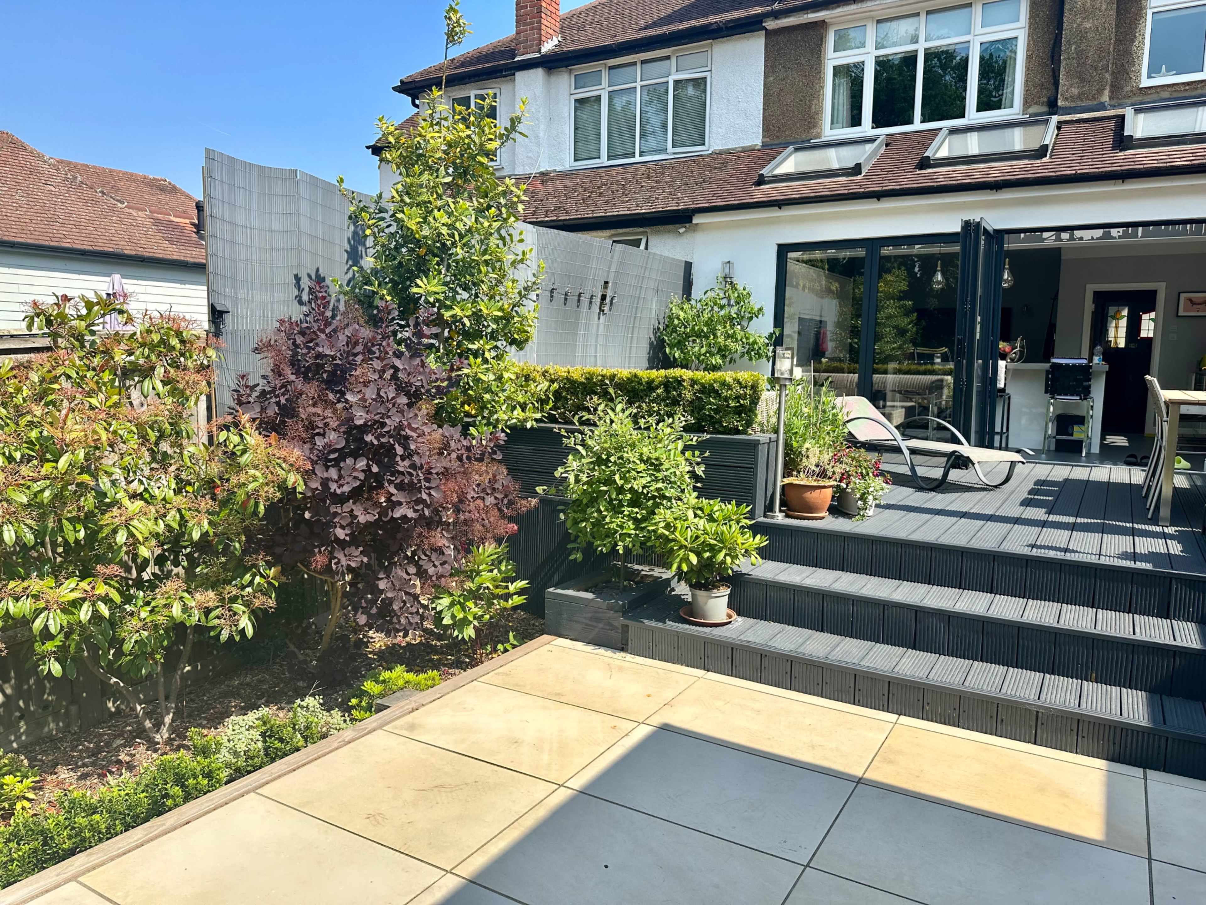 A landscaped backyard features a wooden deck with steps leading up to a modern home, surrounded by various plants and a privacy screen.