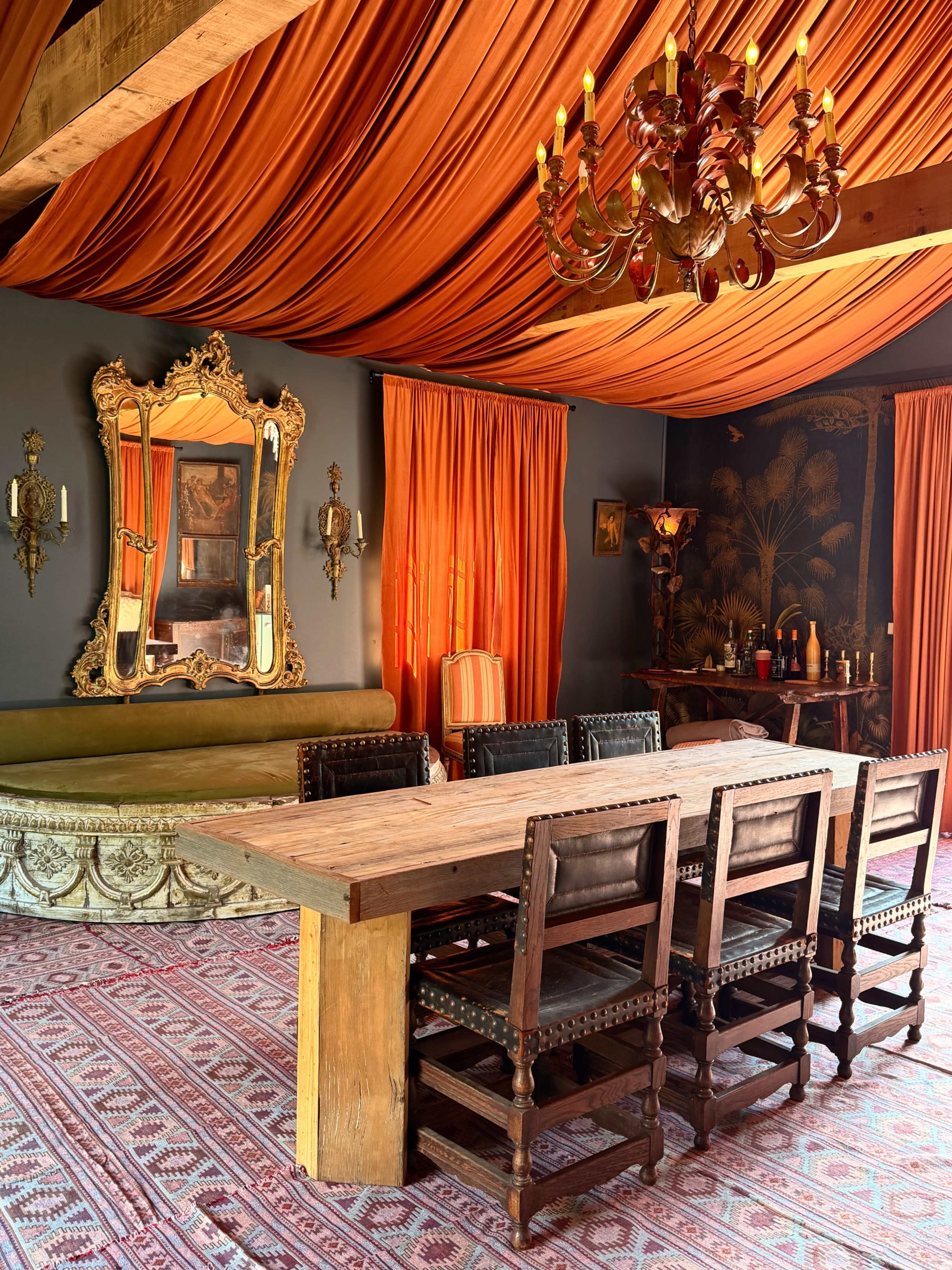 The image shows a dining room with a long wooden table surrounded by eight chairs, orange drapery on the ceiling and windows, and an ornate mirror on the wall.