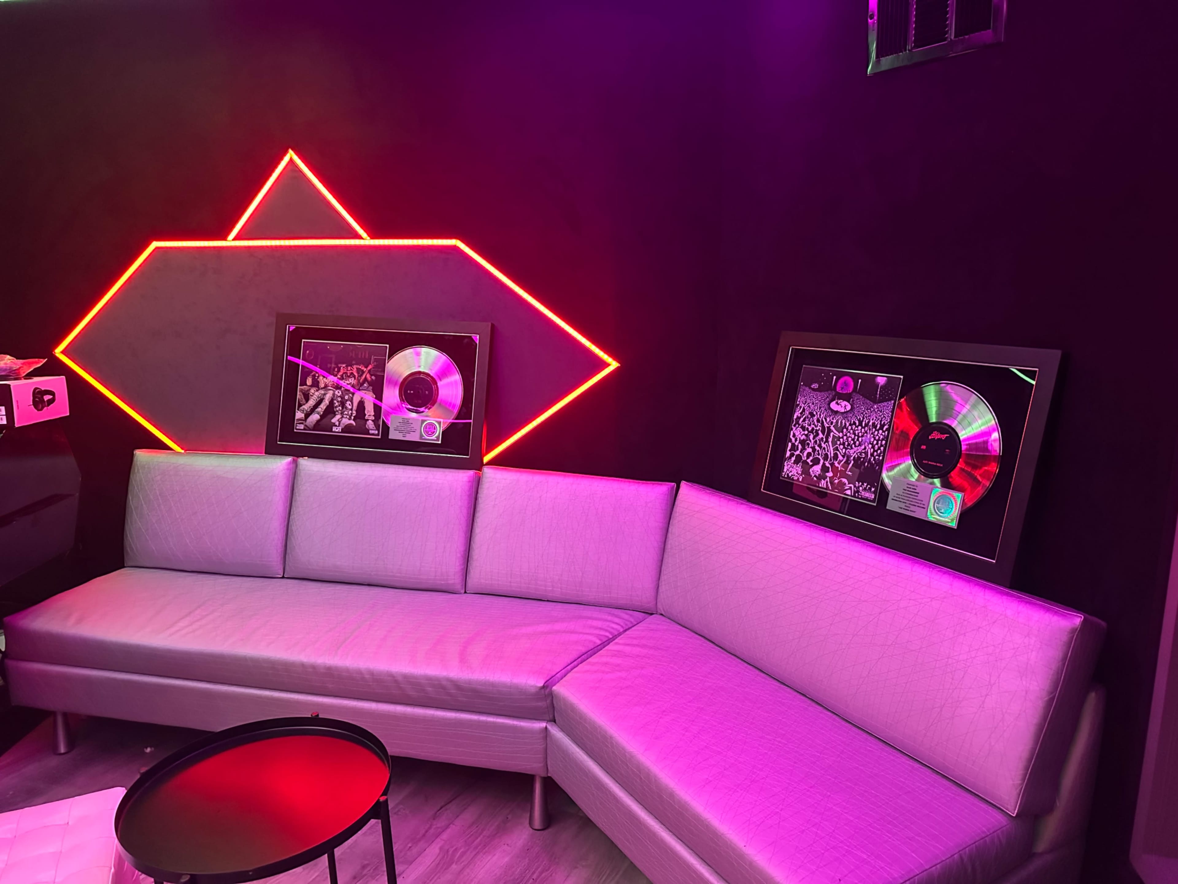 The image shows a modern lounge area with a curved sofa, a small table, and framed music awards displayed on the wall, illuminated by vibrant neon lights.