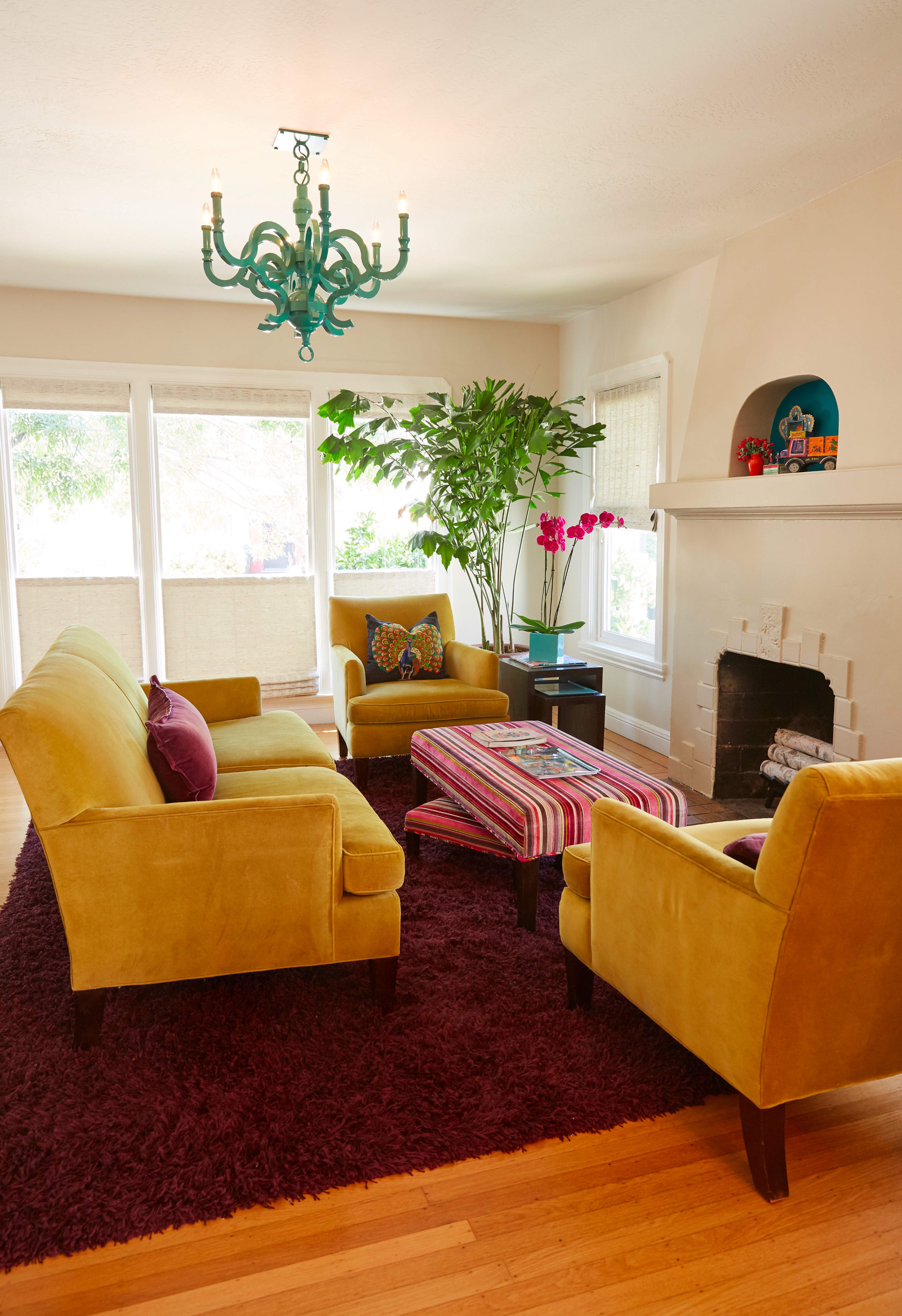 The living room features two yellow chairs and a matching sofa arranged around a striped coffee table, with large windows allowing natural light, a green chandelier overhead, and indoor plants adding to the decor.