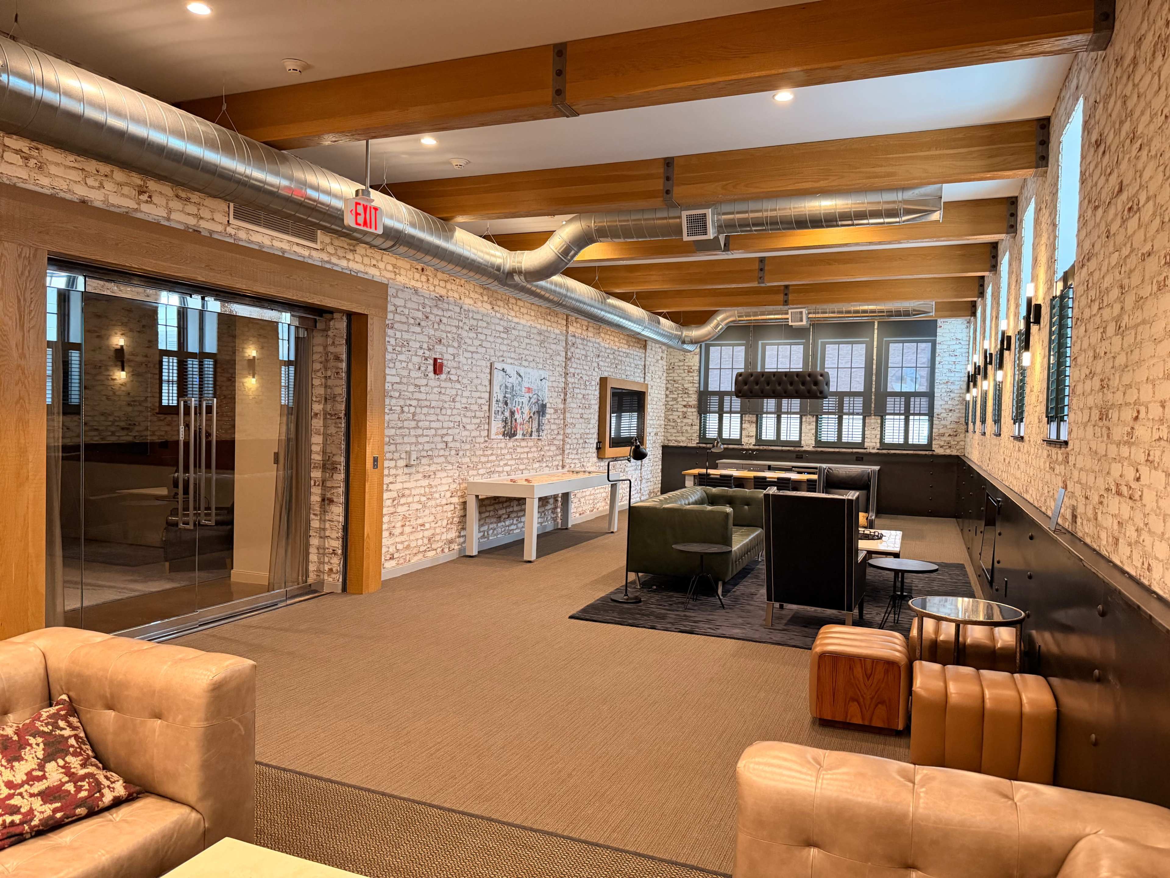 The image shows a well-lit, modern lounge area with exposed brick walls, wooden beams, and a mix of seating including couches and armchairs.