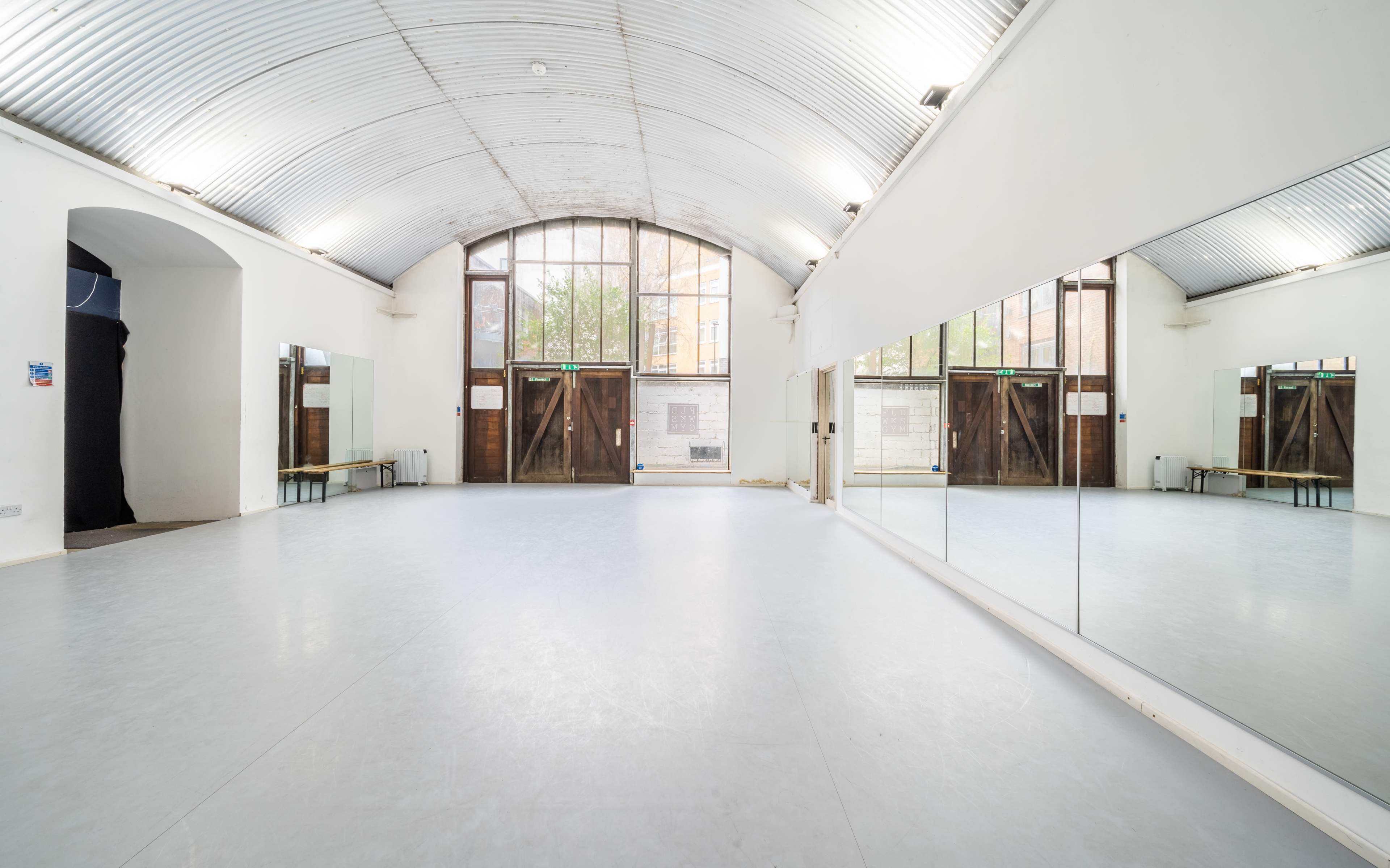 A spacious dance studio features a mirrored wall, vaulted ceiling, and large windows with wooden doors leading outside.