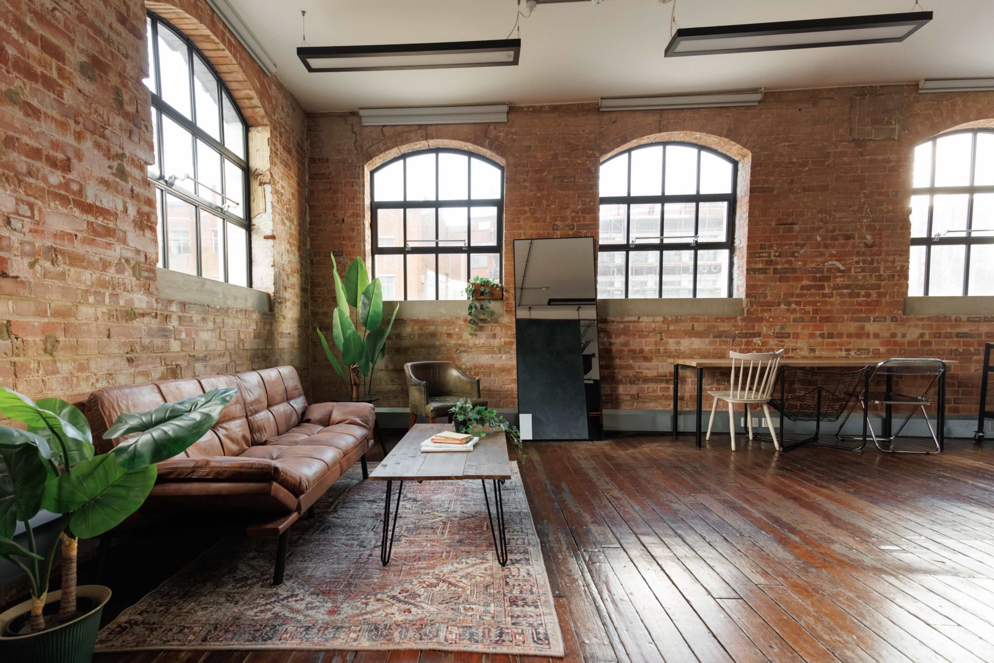 The image shows a spacious room with brick walls, large windows, a brown leather sofa, a coffee table, and a dining area with wooden chairs.