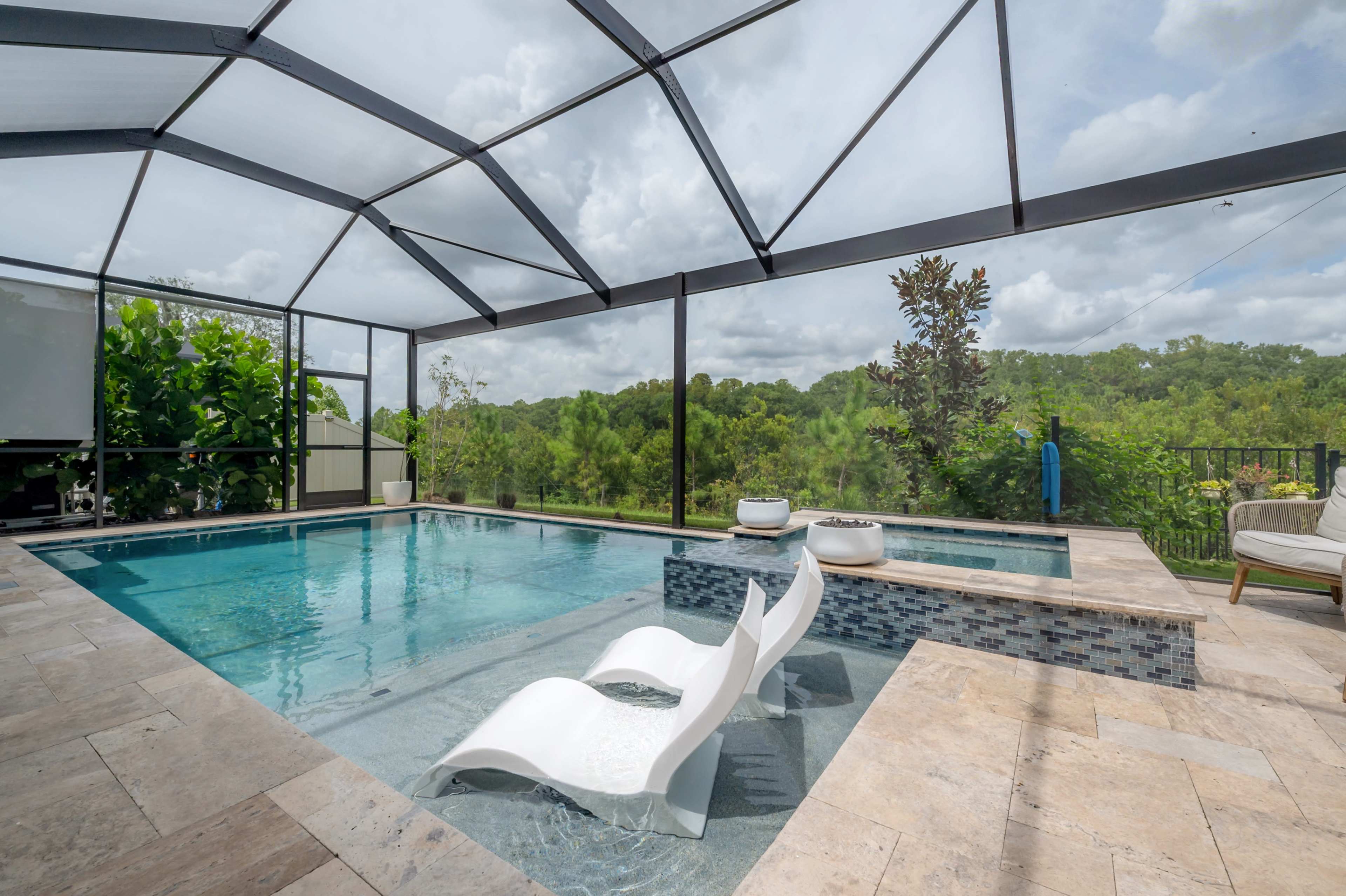 The image shows a modern pool area with two white lounge chairs, surrounded by a landscaped backdrop of greenery, all under a screened enclosure.