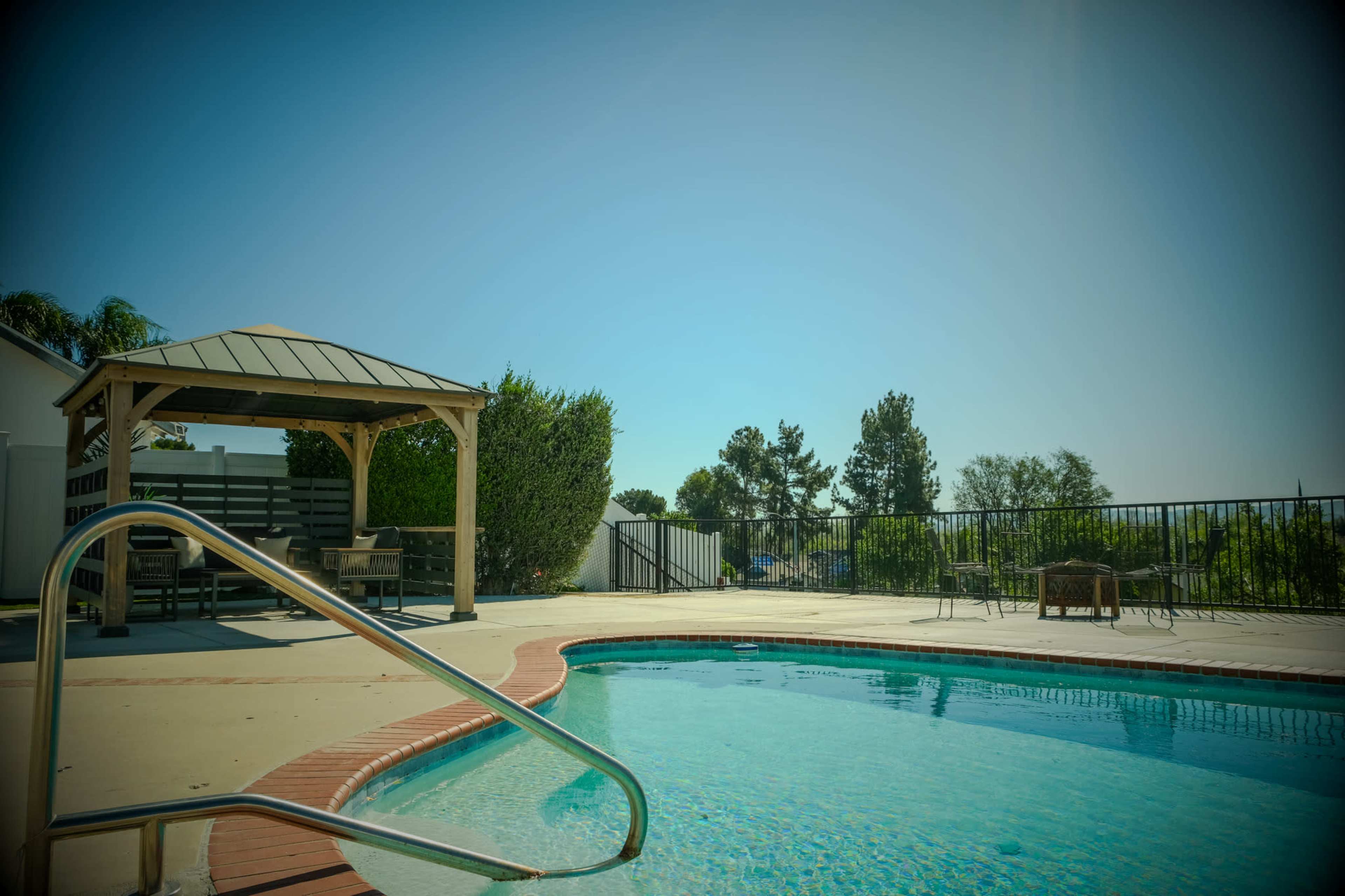 The image shows a swimming pool with a pergola and seating area in a landscaped backyard.