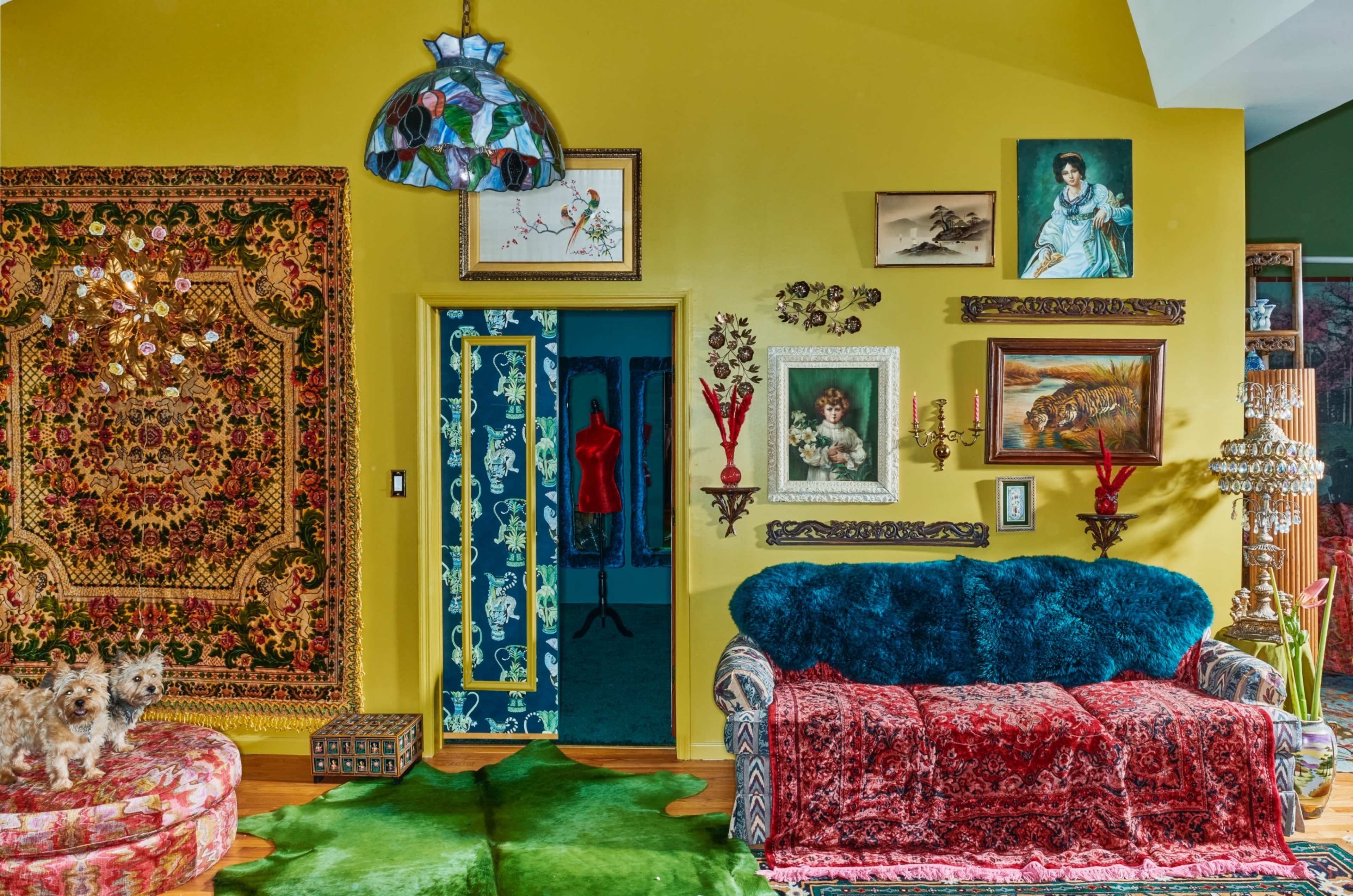 The room features a vibrant yellow wall decorated with various frames and art, a plush couch covered in red fabric, and two small dogs on a green carpet.