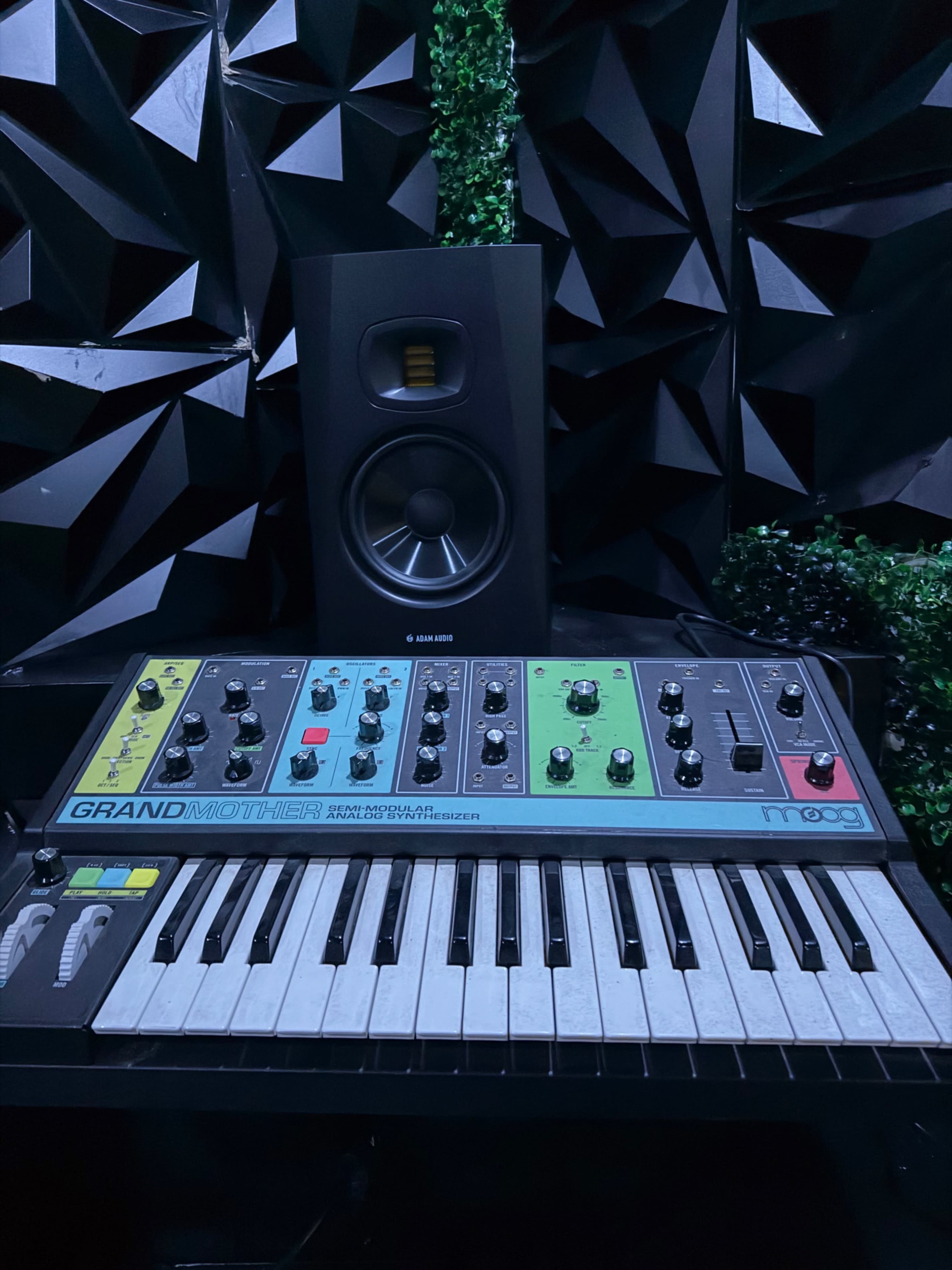 The image shows a synthesizer called "GRANDMOTHER" positioned in front of a black speaker, with a textured wall of triangular panels in the background.