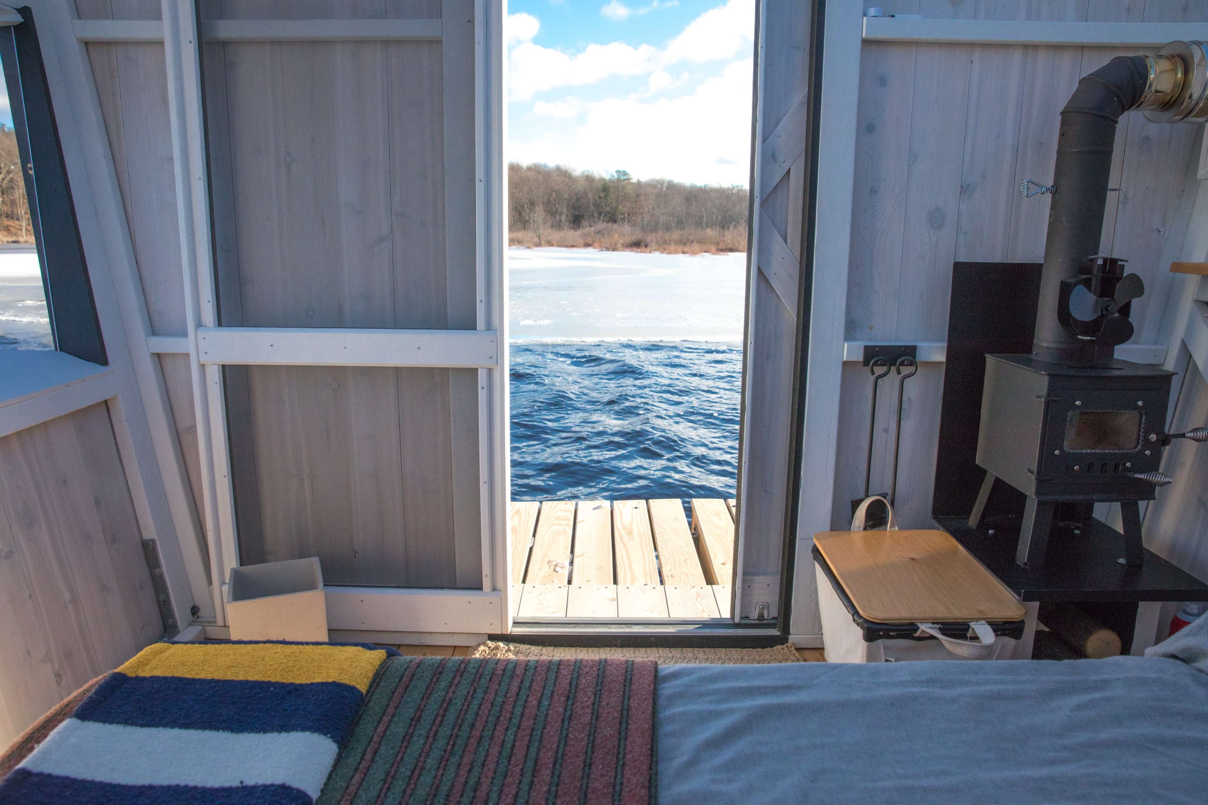 A cozy, rustic interior view of a small cabin with a wood stove, leading out to a frozen lake with wooden decking.