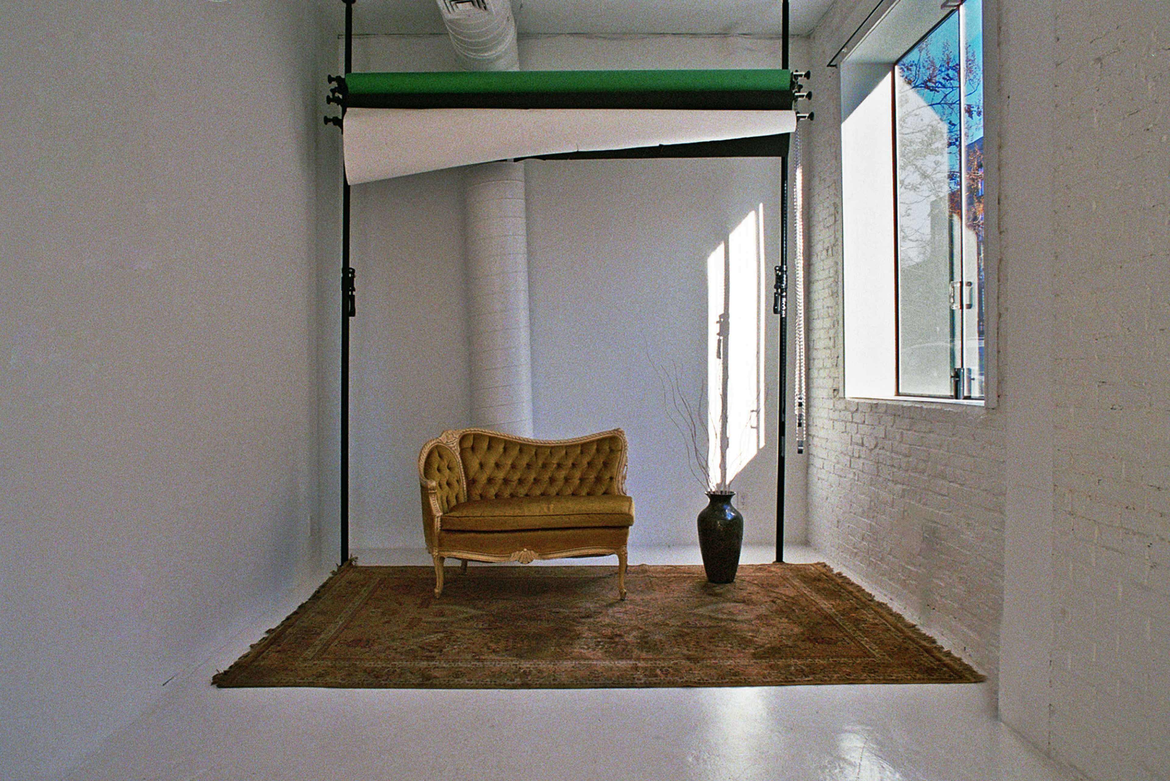 The image shows a minimalist photography studio with a vintage yellow sofa, a vase, and large windows letting in natural light.