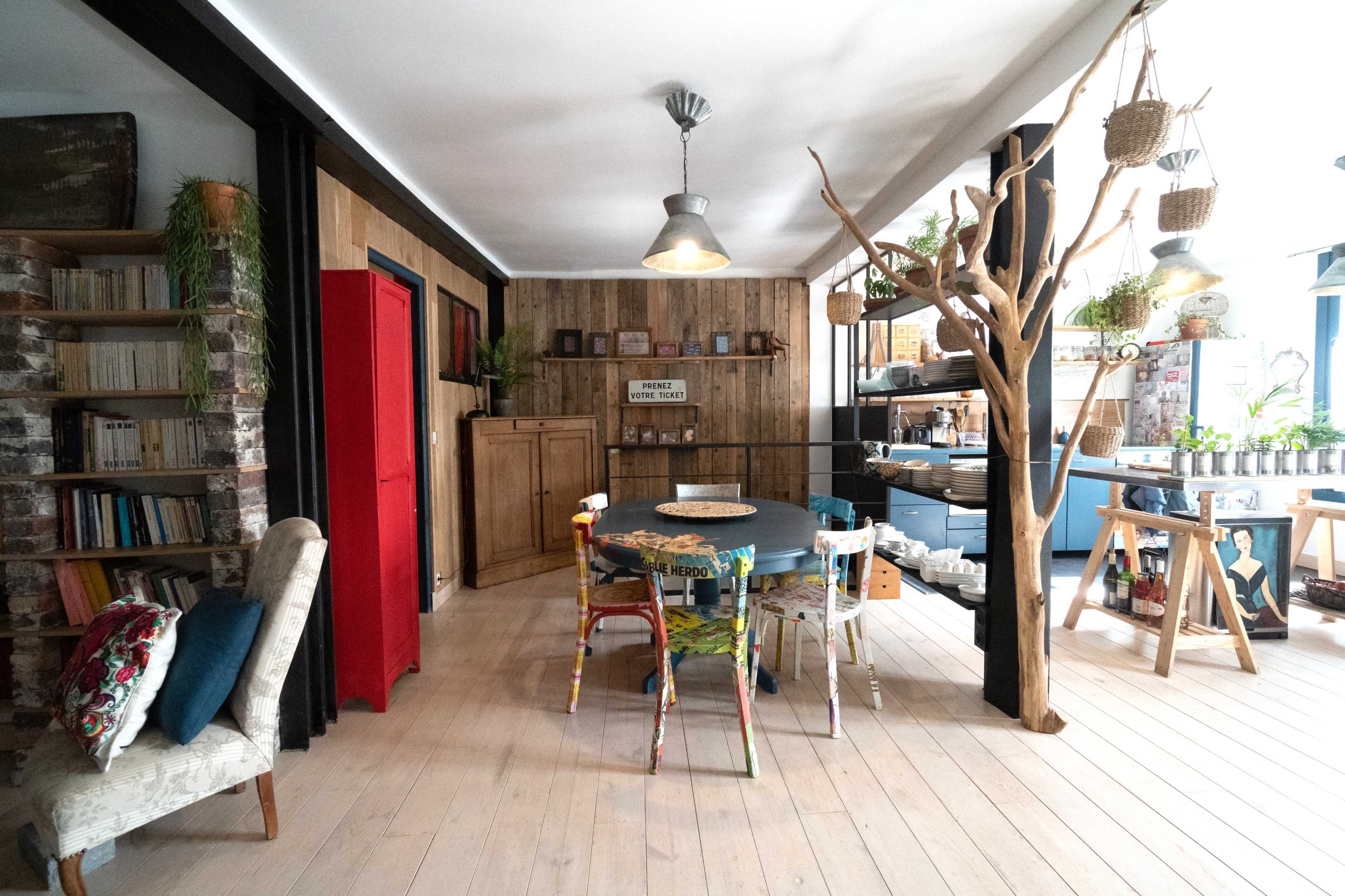 A cozy interior space featuring a dining table surrounded by colorful chairs, a wooden bookshelf, and various plants integrated into the rustic decor.