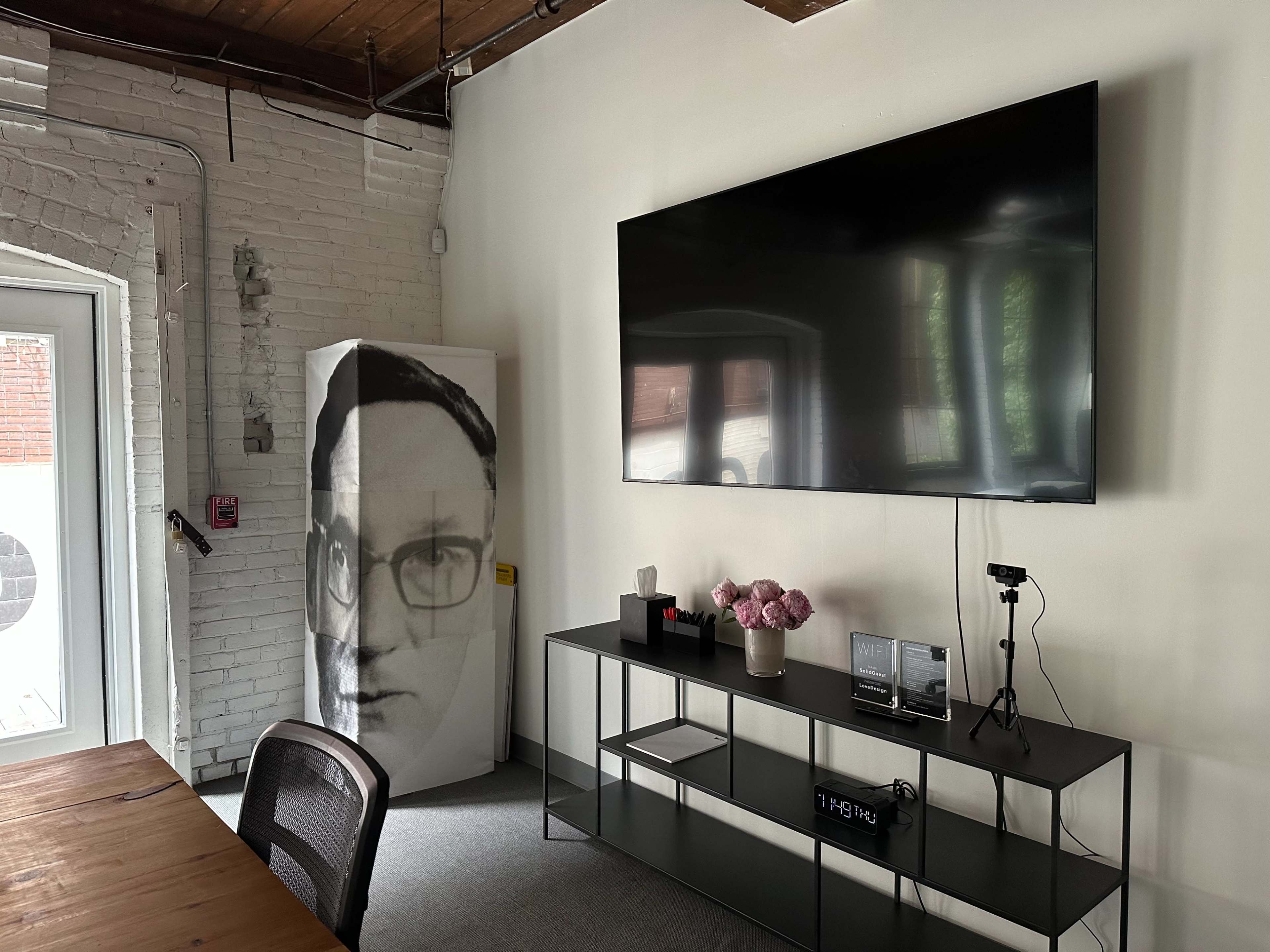 A modern office space featuring a large television mounted on the wall, a black metal shelf holding decorative items, and a refrigerator with a graphic portrait on its front.