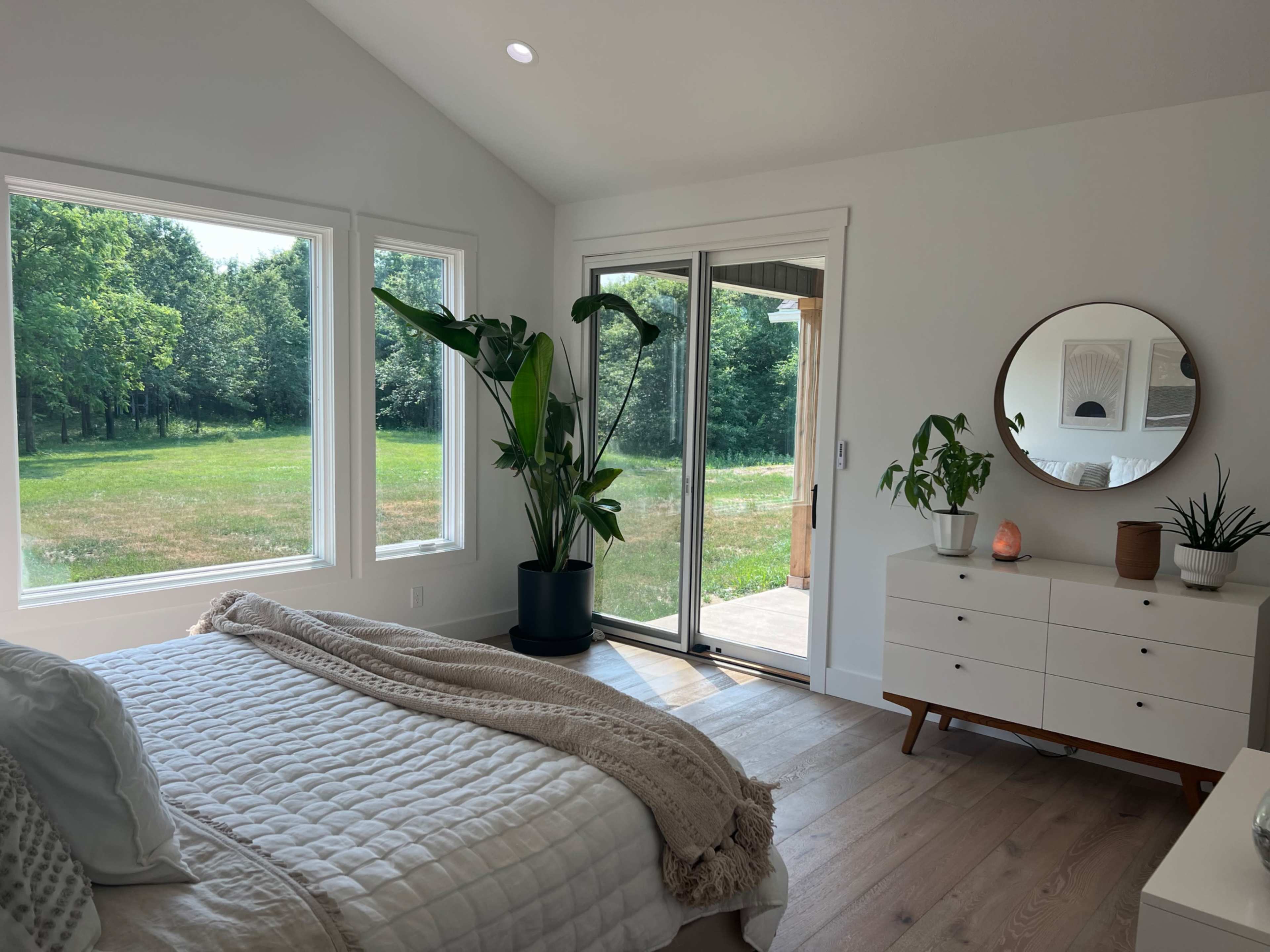 A bright bedroom features large windows with views of greenery, a sliding glass door, a bed with a textured blanket, and a dresser adorned with plants and a mirror.