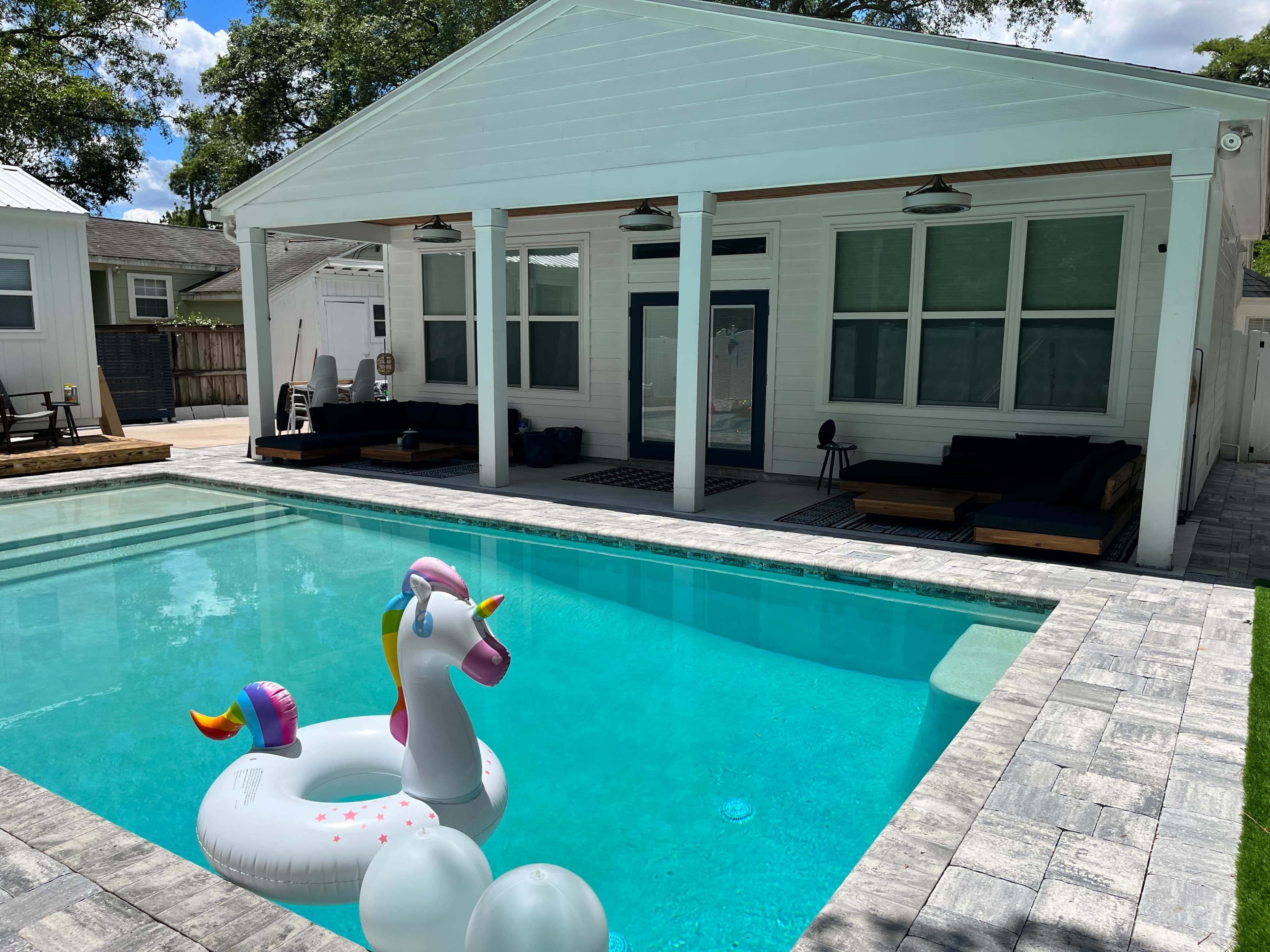 A backyard pool area with a unicorn-shaped inflatable float in the water and a modern white house in the background.