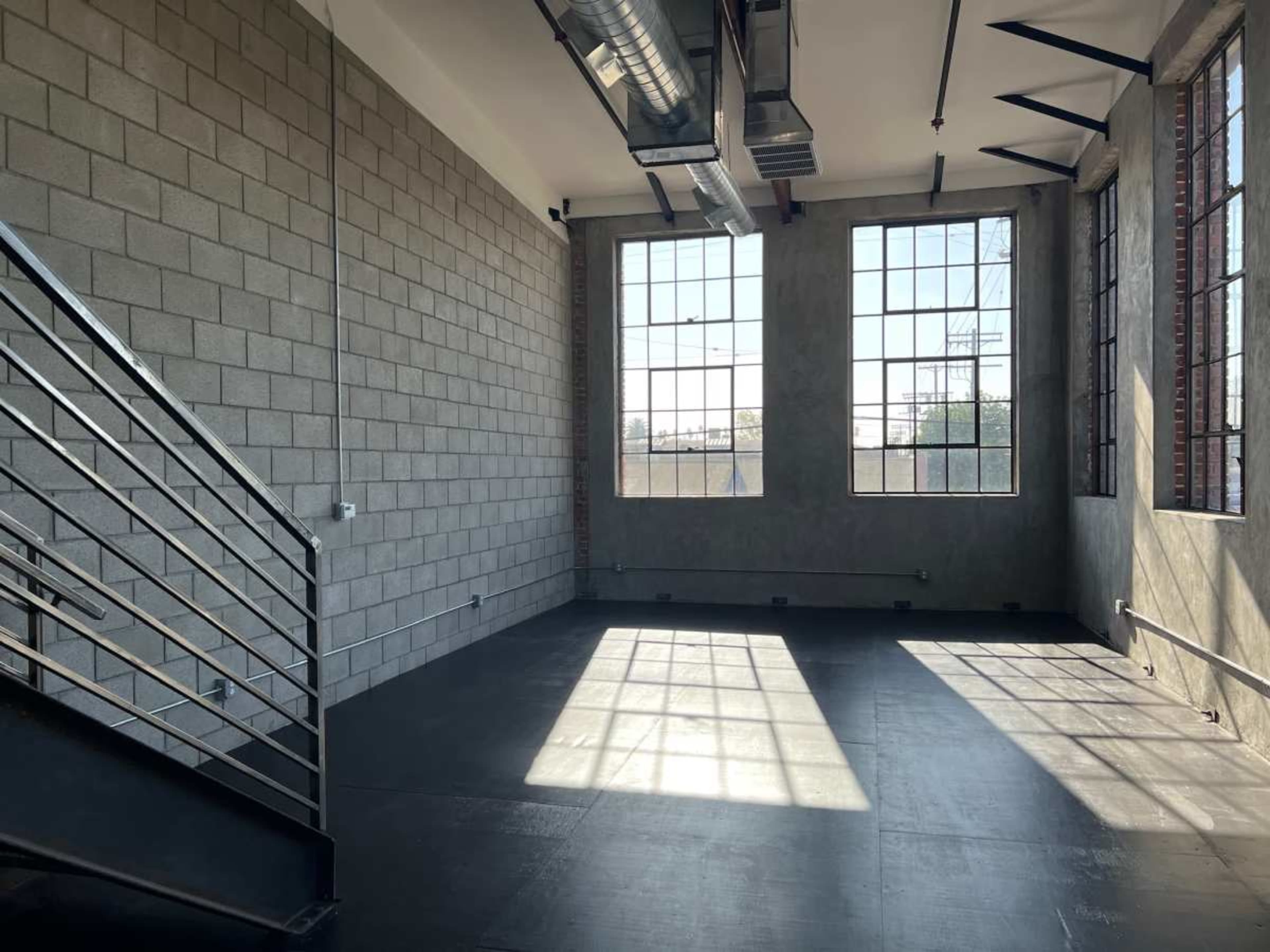 The image shows an industrial-style interior space with large windows, concrete walls, and a staircase leading to a lower level.