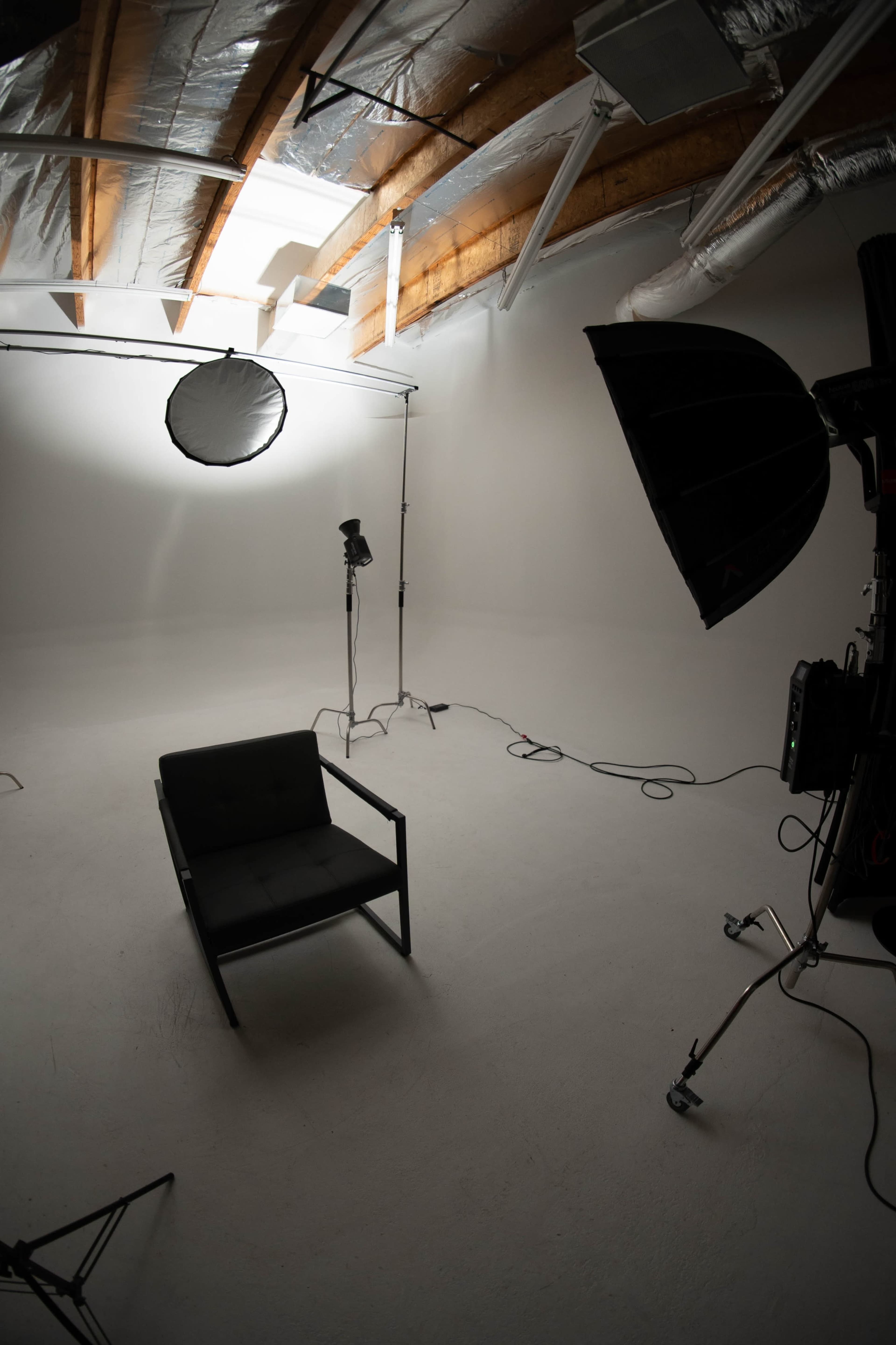 The image shows a minimalistic photography studio with a black chair, lighting equipment, and a white backdrop.