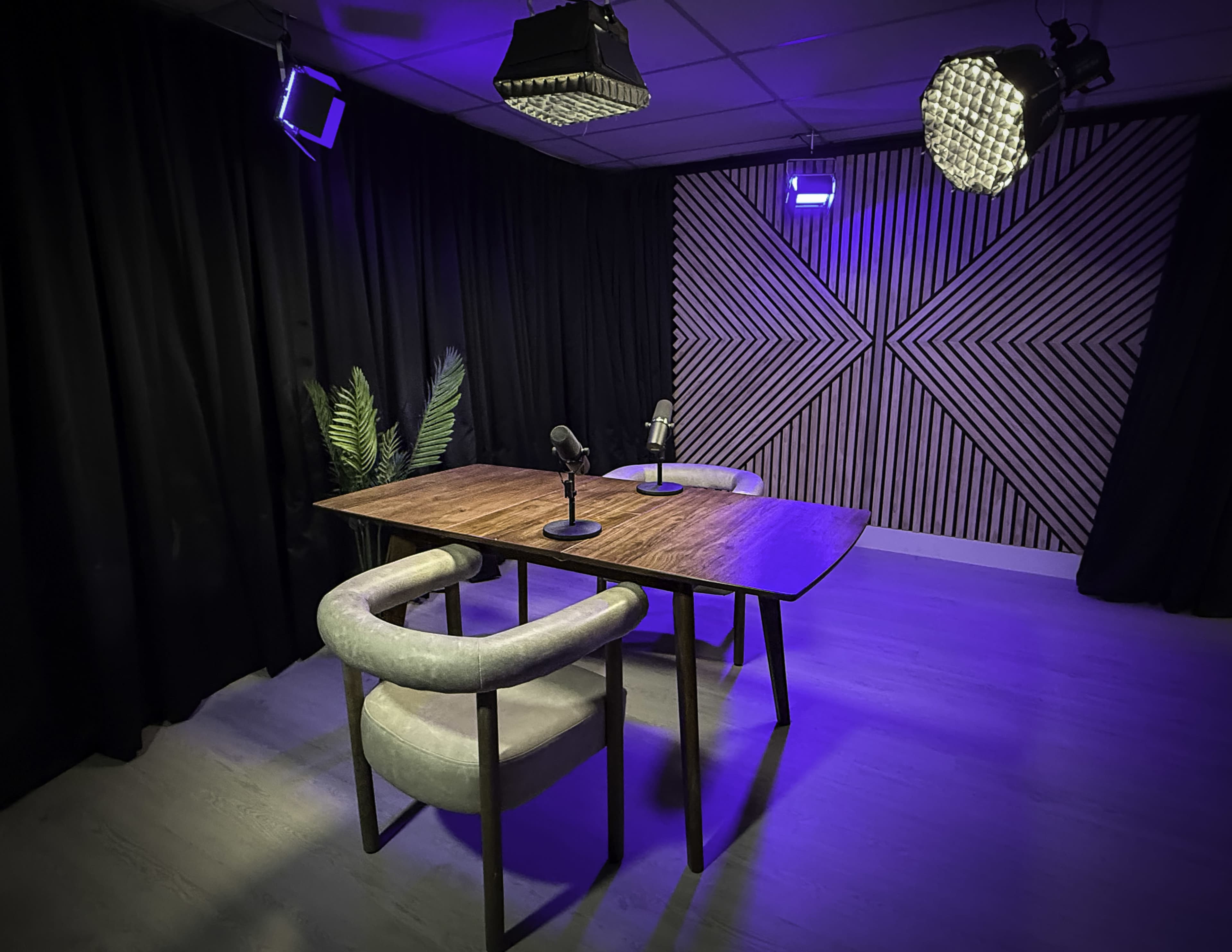 A modern recording studio features a wooden table with two microphones, surrounded by dark curtains and patterned wooden walls, illuminated by bright lights.