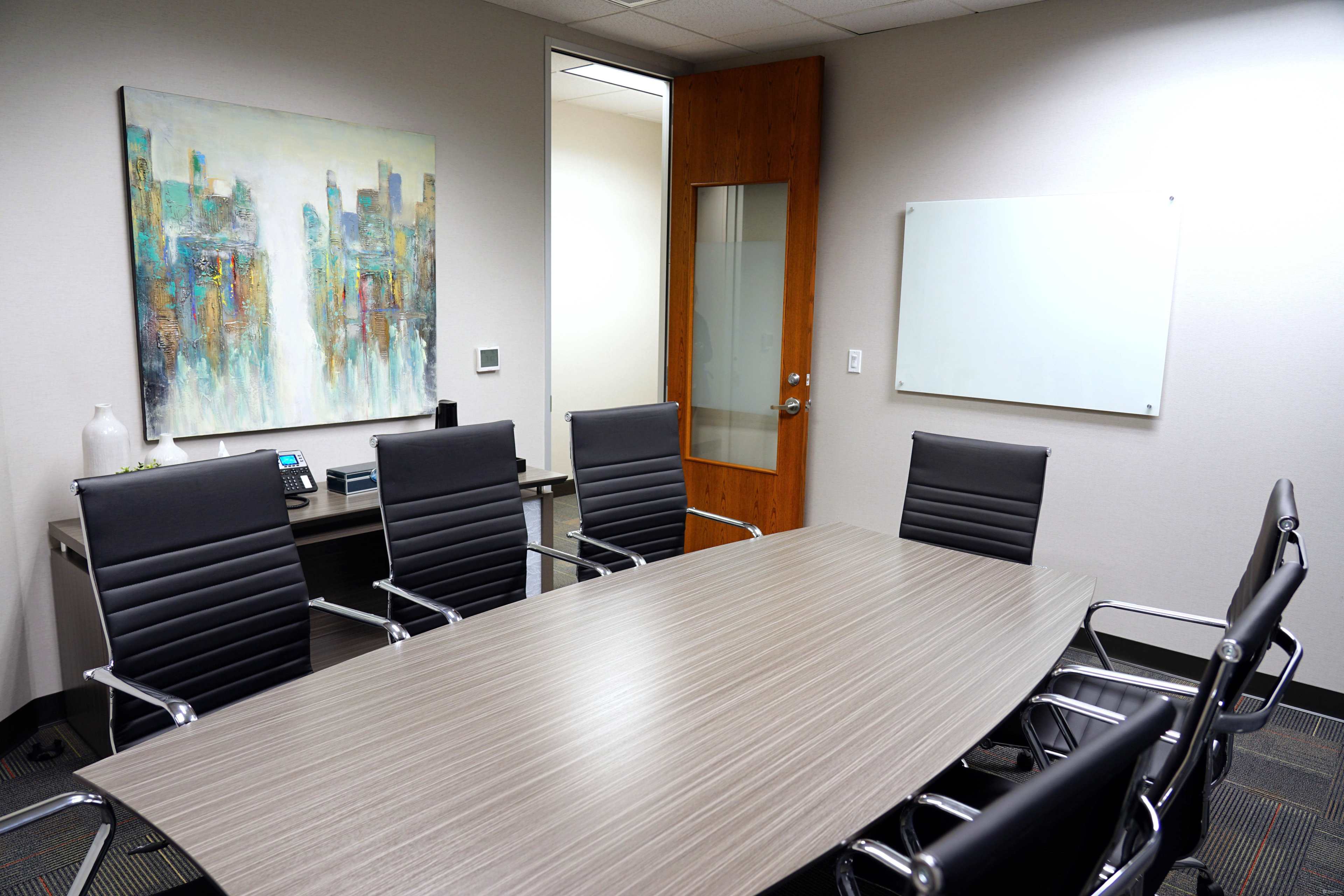 A modern conference room features a large oval table surrounded by eight black leather chairs, with a colorful abstract painting on the wall and a whiteboard nearby.