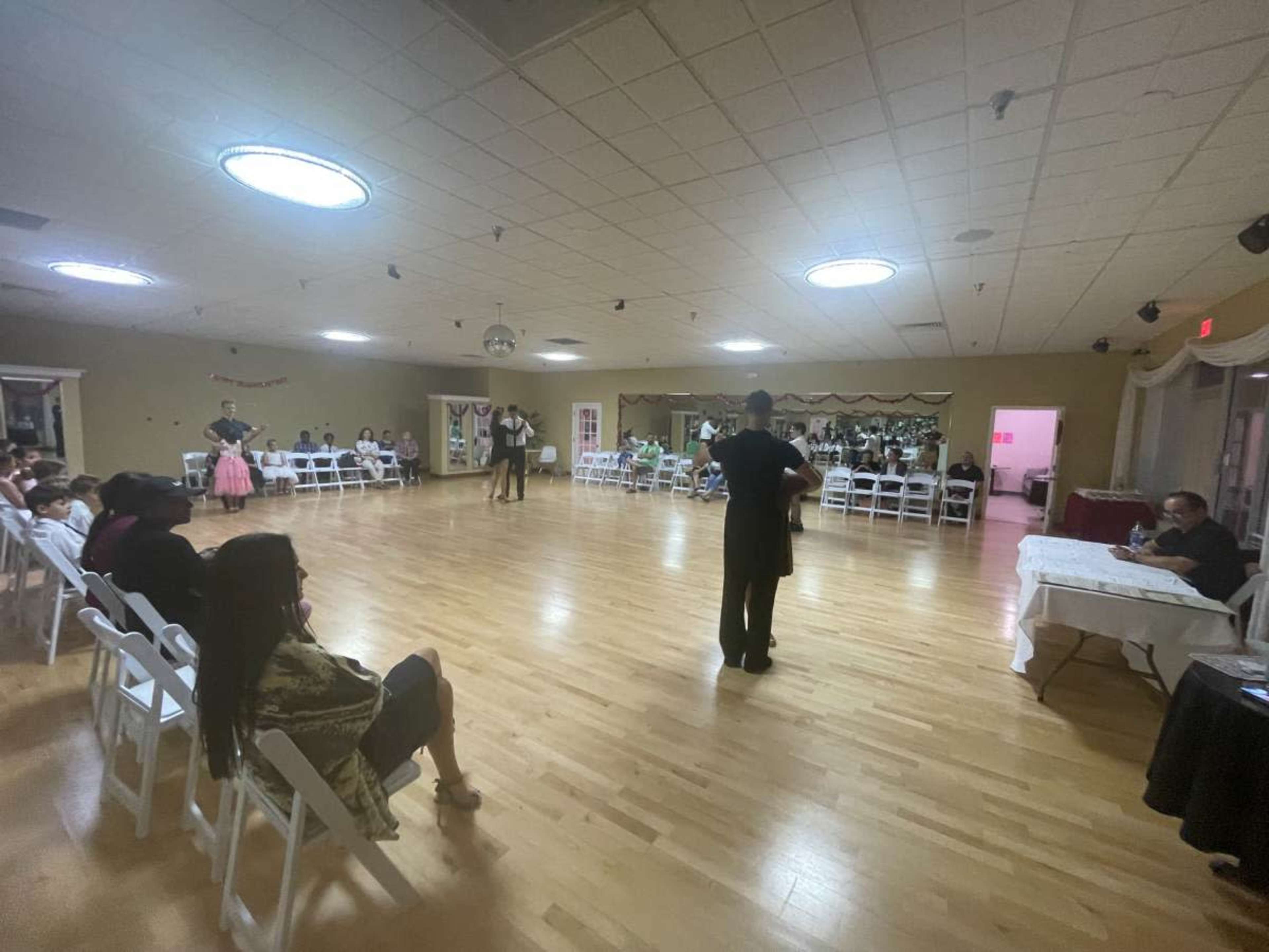 A formal gathering is taking place in a spacious hall, with attendees seated in rows while a couple dances in the center.