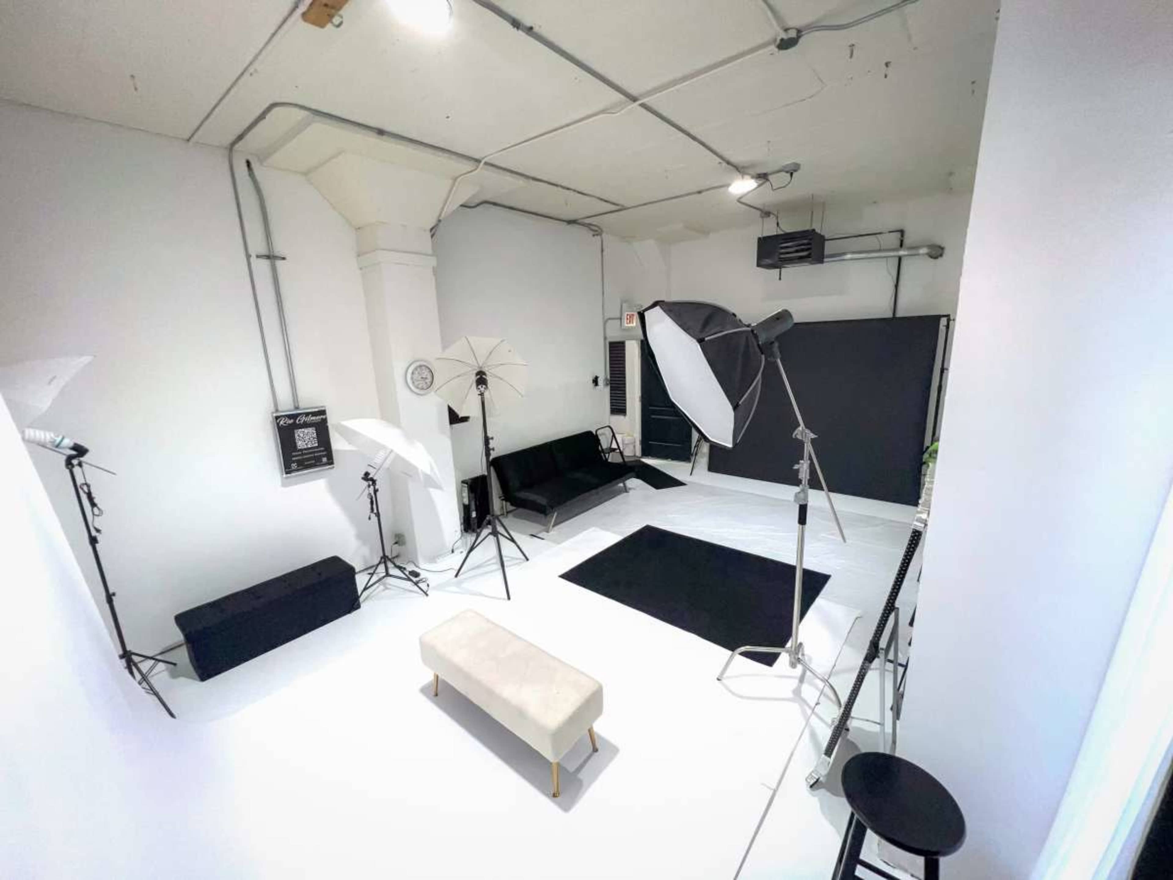 The image shows a photography studio with a black sofa, a white bench, a large backdrop, and various lighting equipment arranged in a bright, minimalist space.