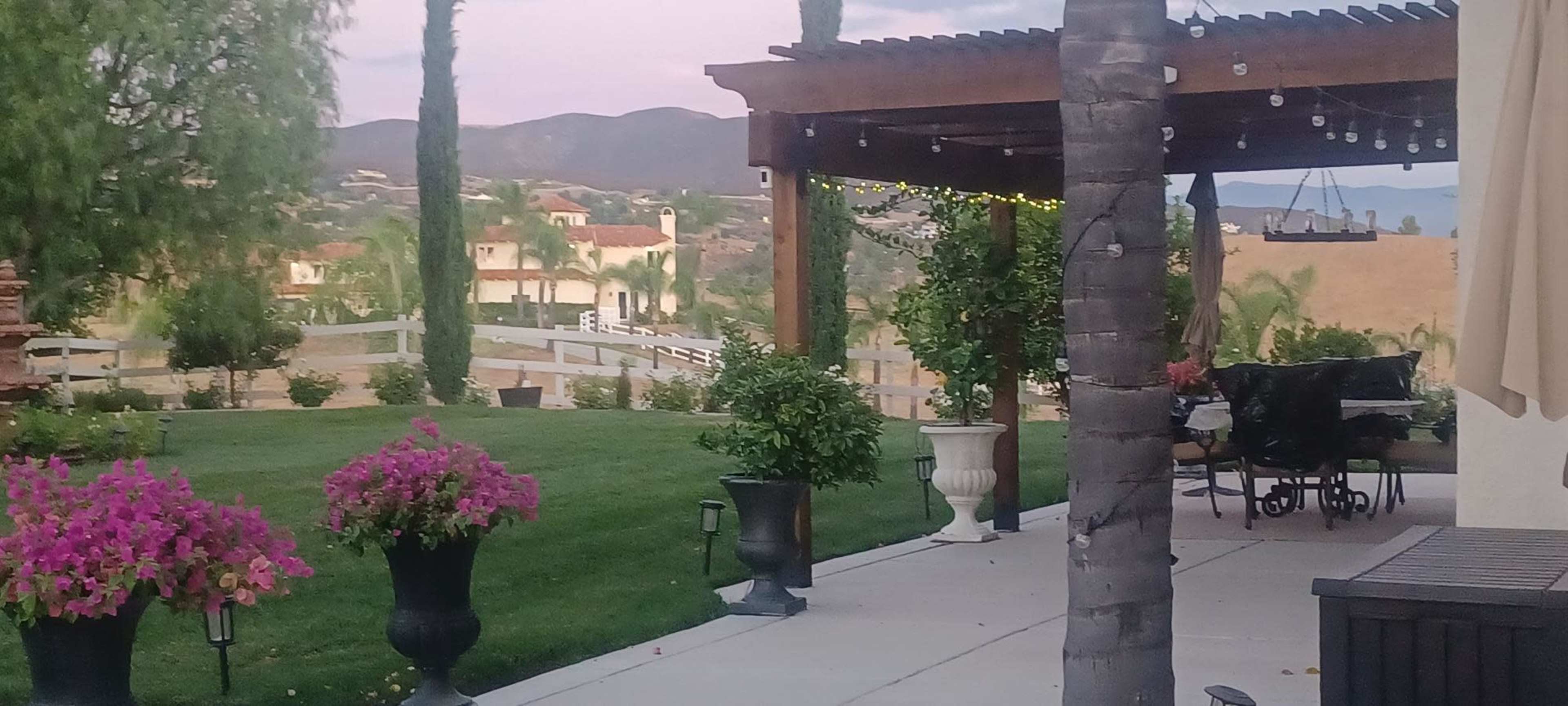 The image shows a landscaped backyard with potted plants, a patio area, and distant rolling hills.
