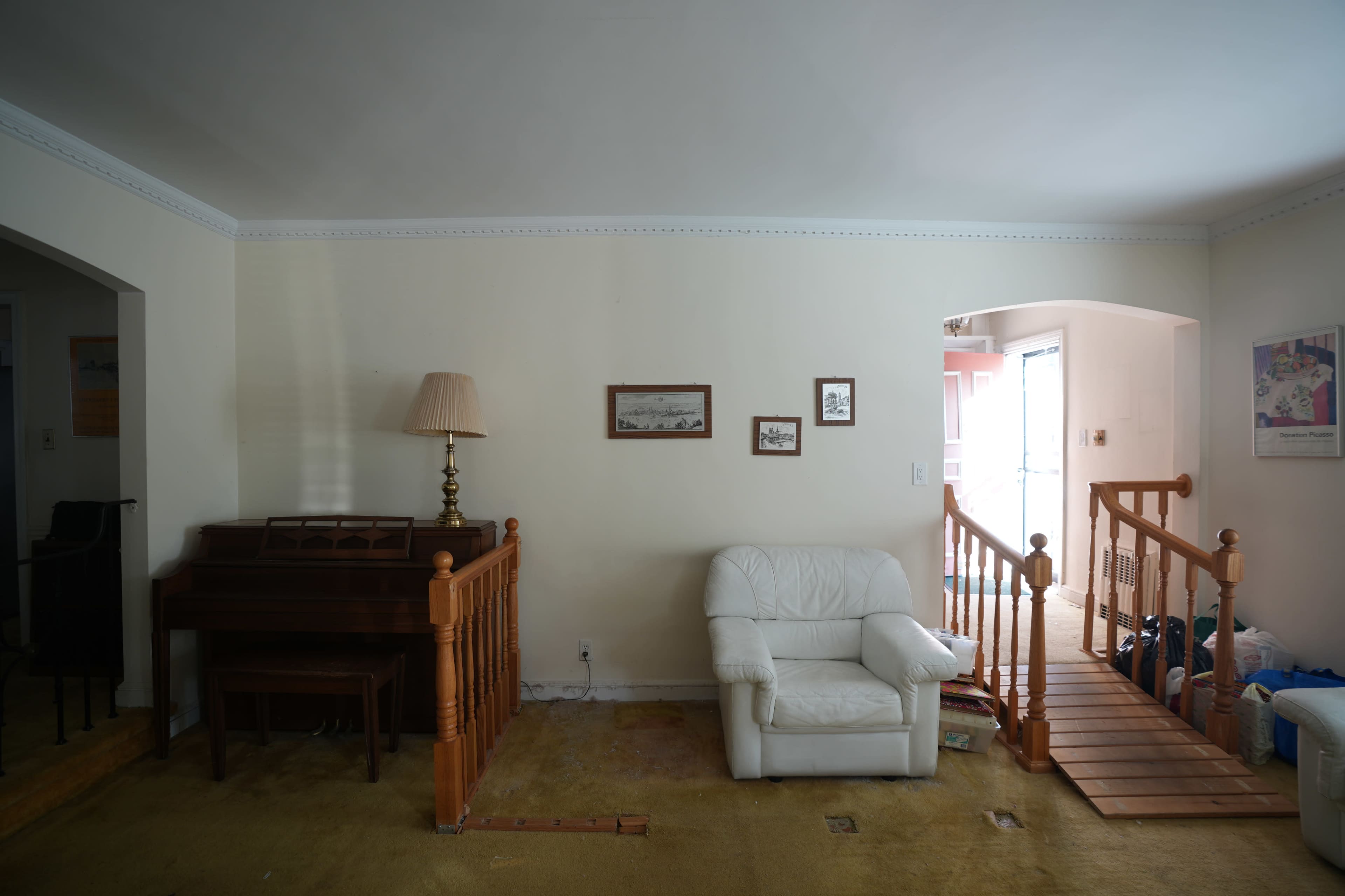 The image shows a living room with a white armchair, a piano, and a staircase leading to another room, along with bare walls and carpet.