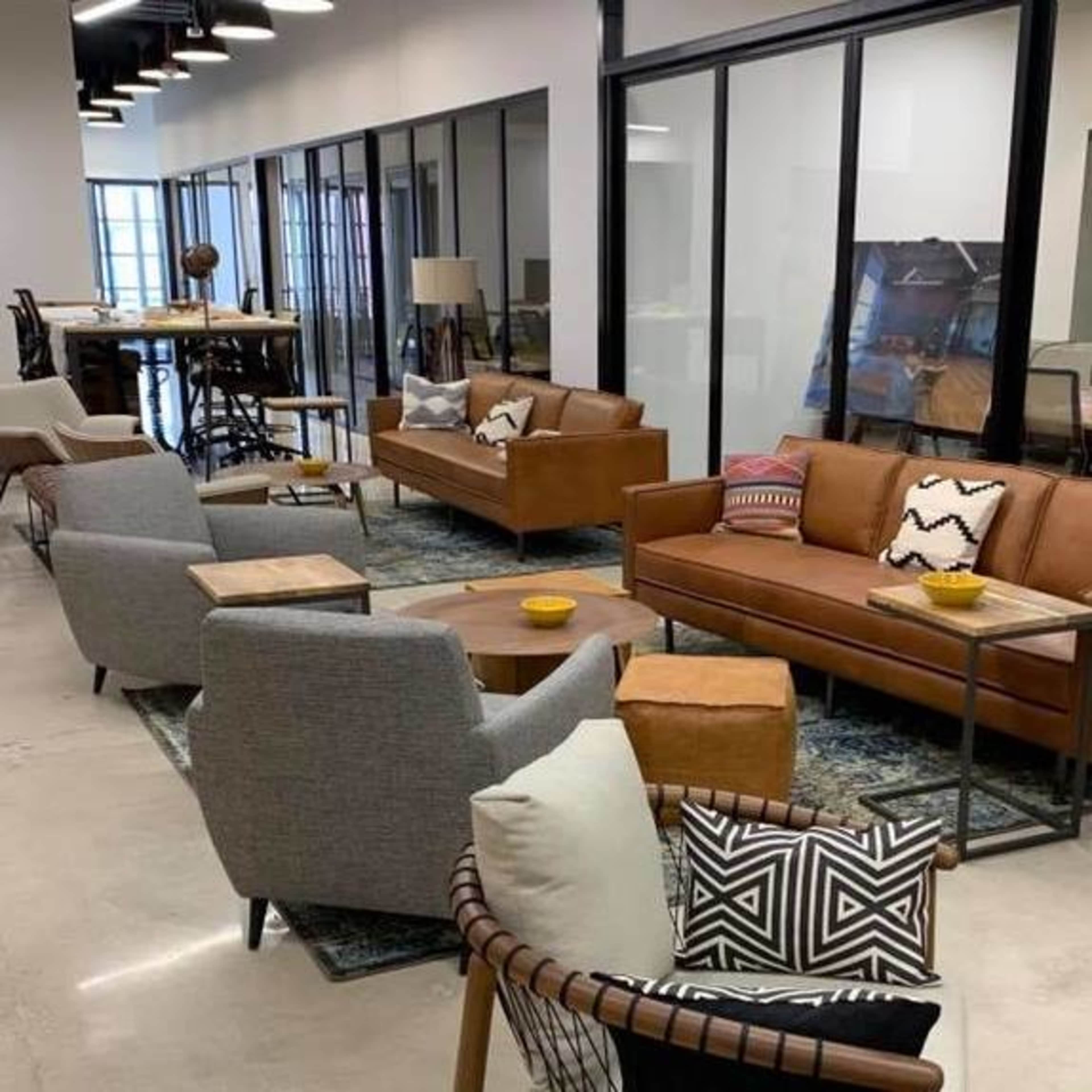 The image shows a modern office space featuring a seating area with brown leather sofas, gray armchairs, and decorative cushions, surrounded by glass-enclosed meeting rooms.