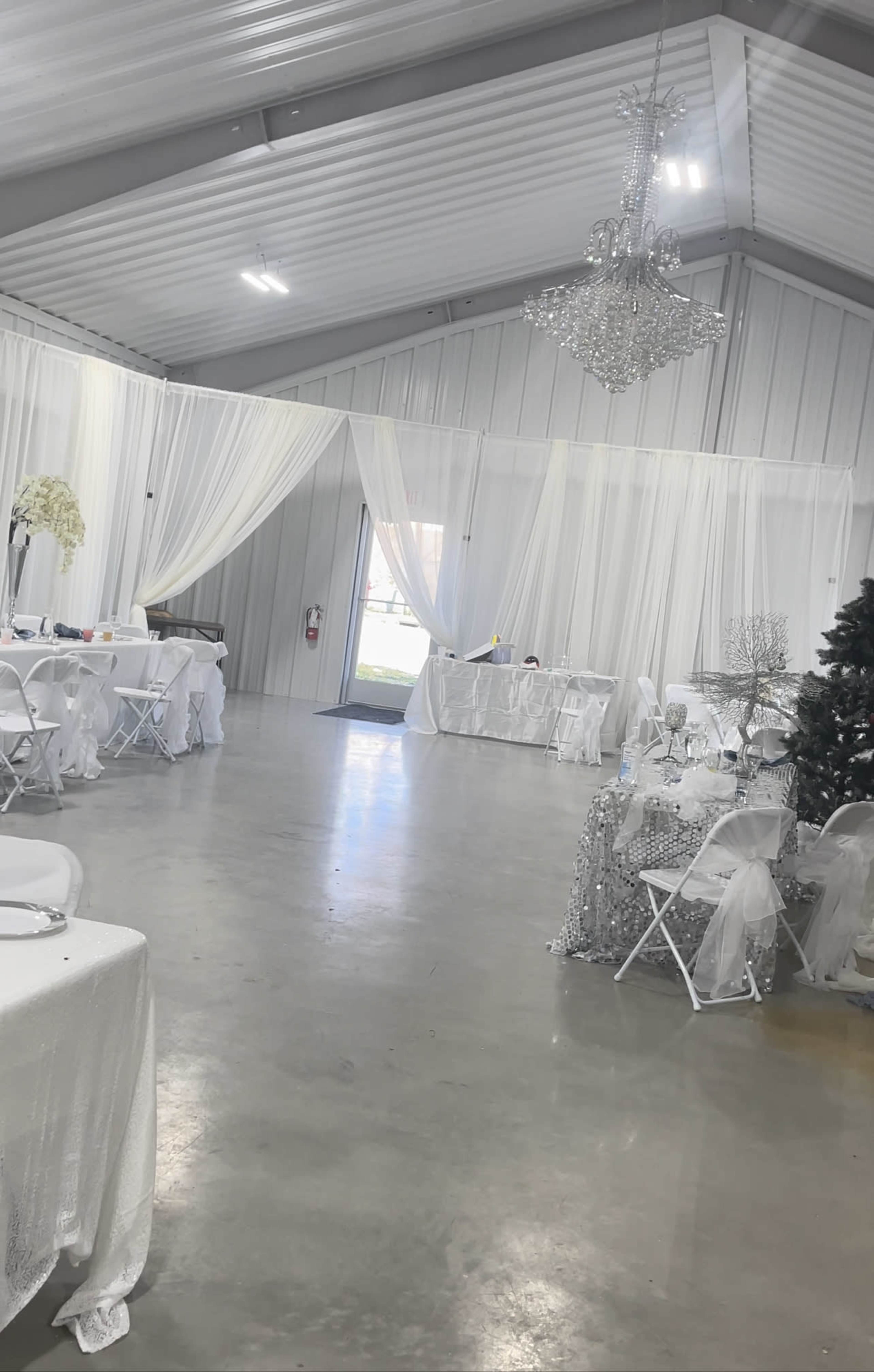 A spacious indoor venue decorated with white drapes and tables, featuring a chandelier and a Christmas tree.