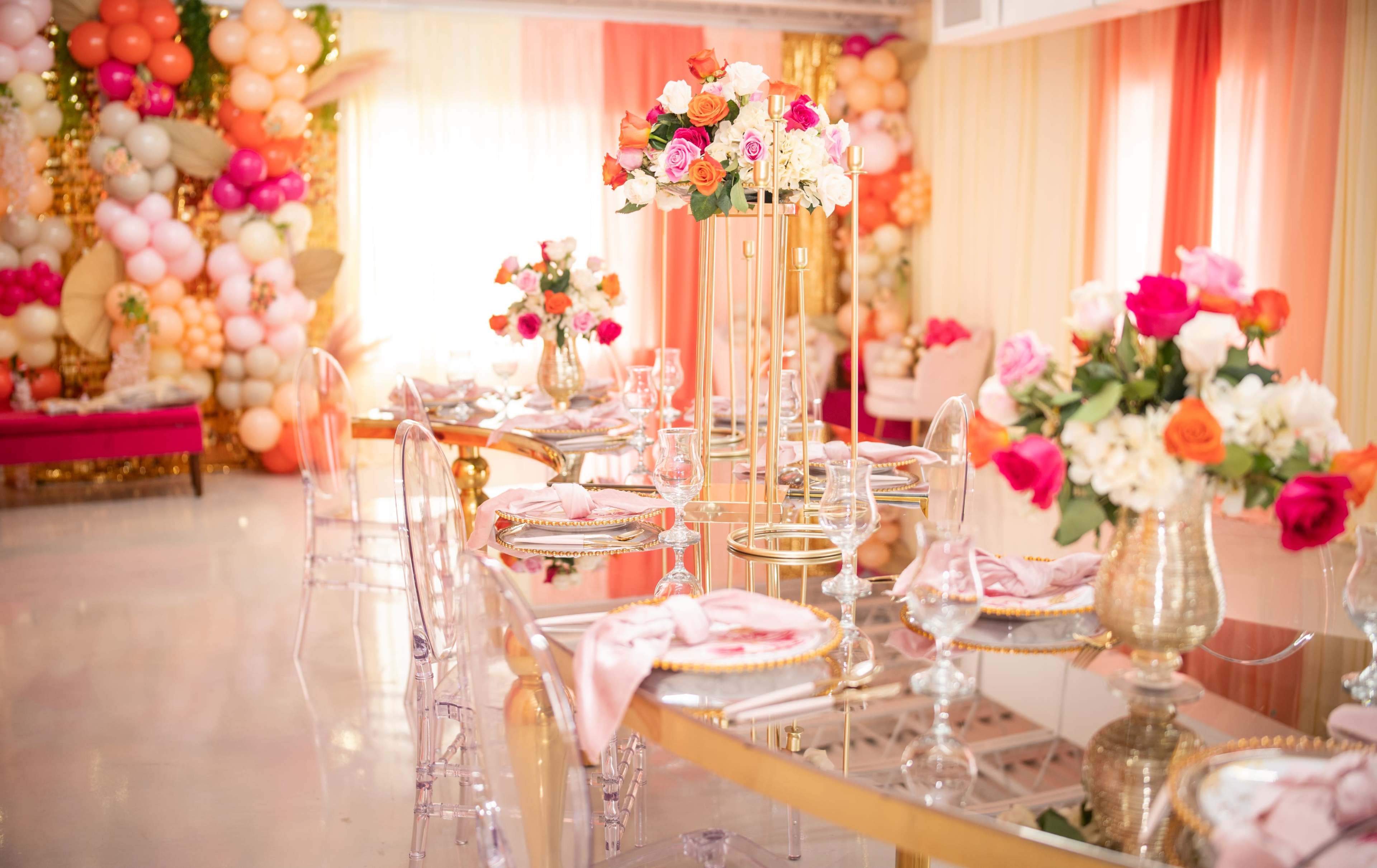 A dining area is set with elegant tables adorned with floral centerpieces and a backdrop of colorful balloons.