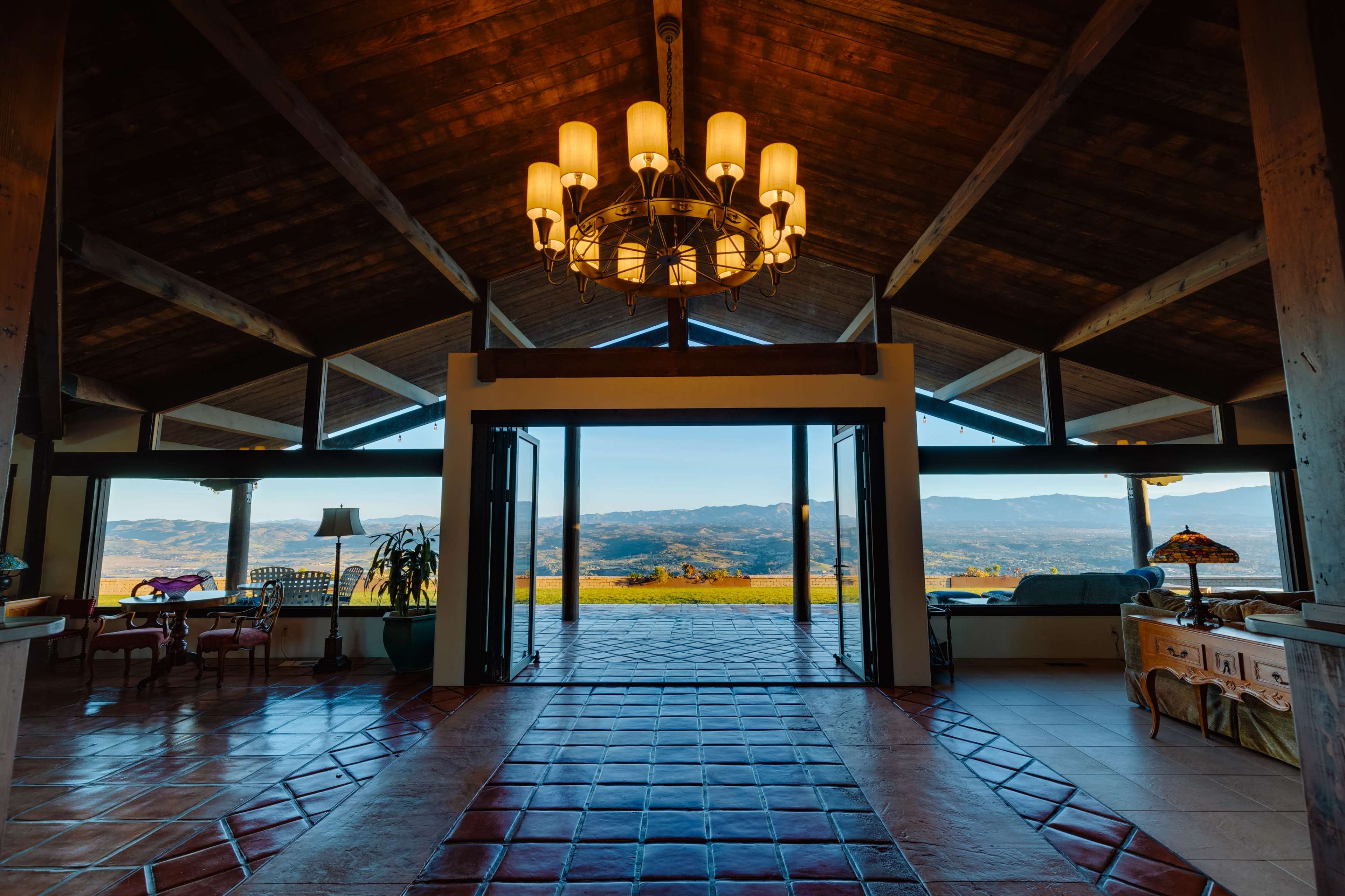 The image shows a spacious interior of a house with large windows overlooking a mountainous landscape, featuring wooden beams and a chandelier.