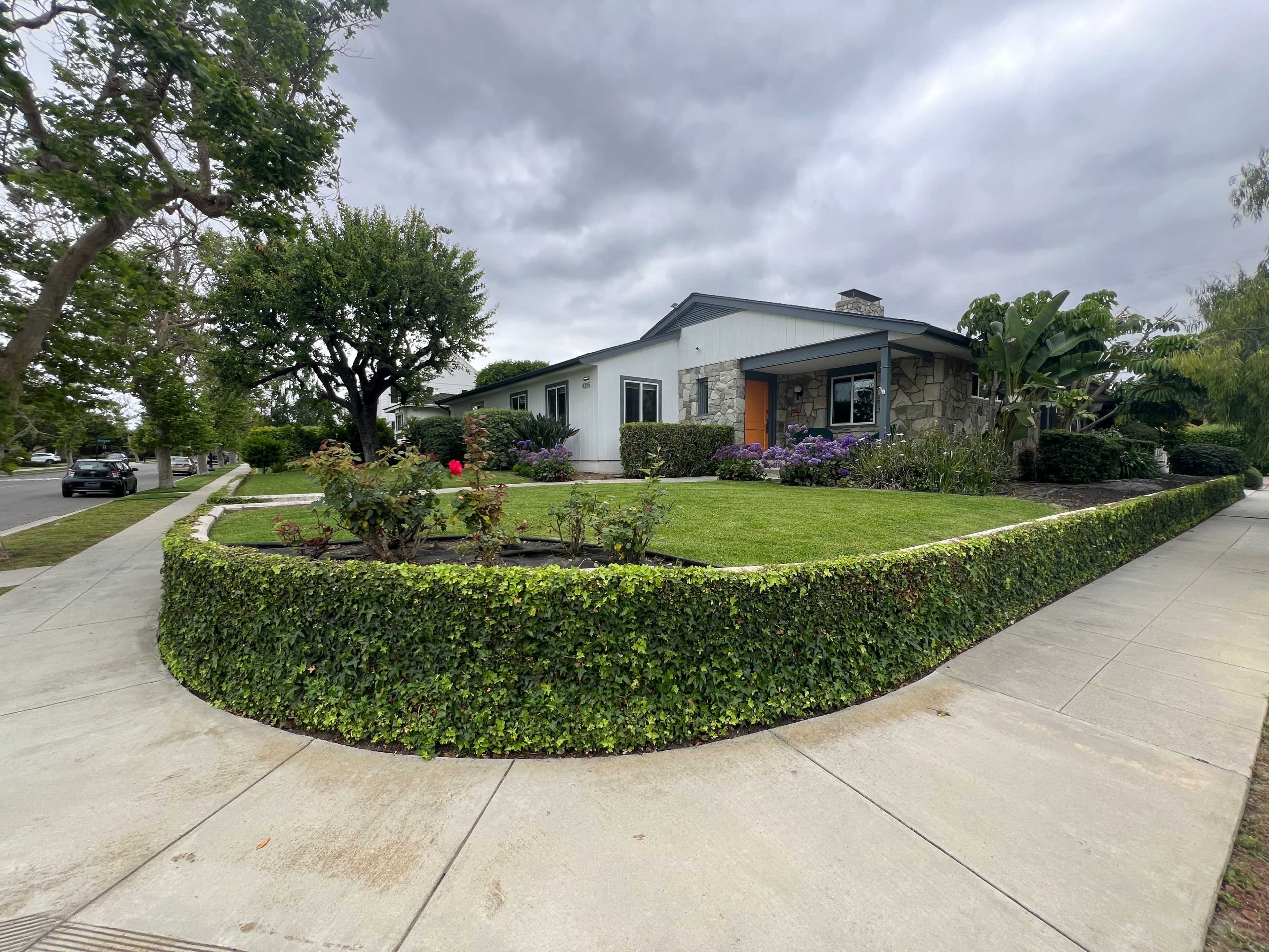 A single-story house with a stone facade and well-maintained lawn is framed by a landscaped garden and curved hedges.