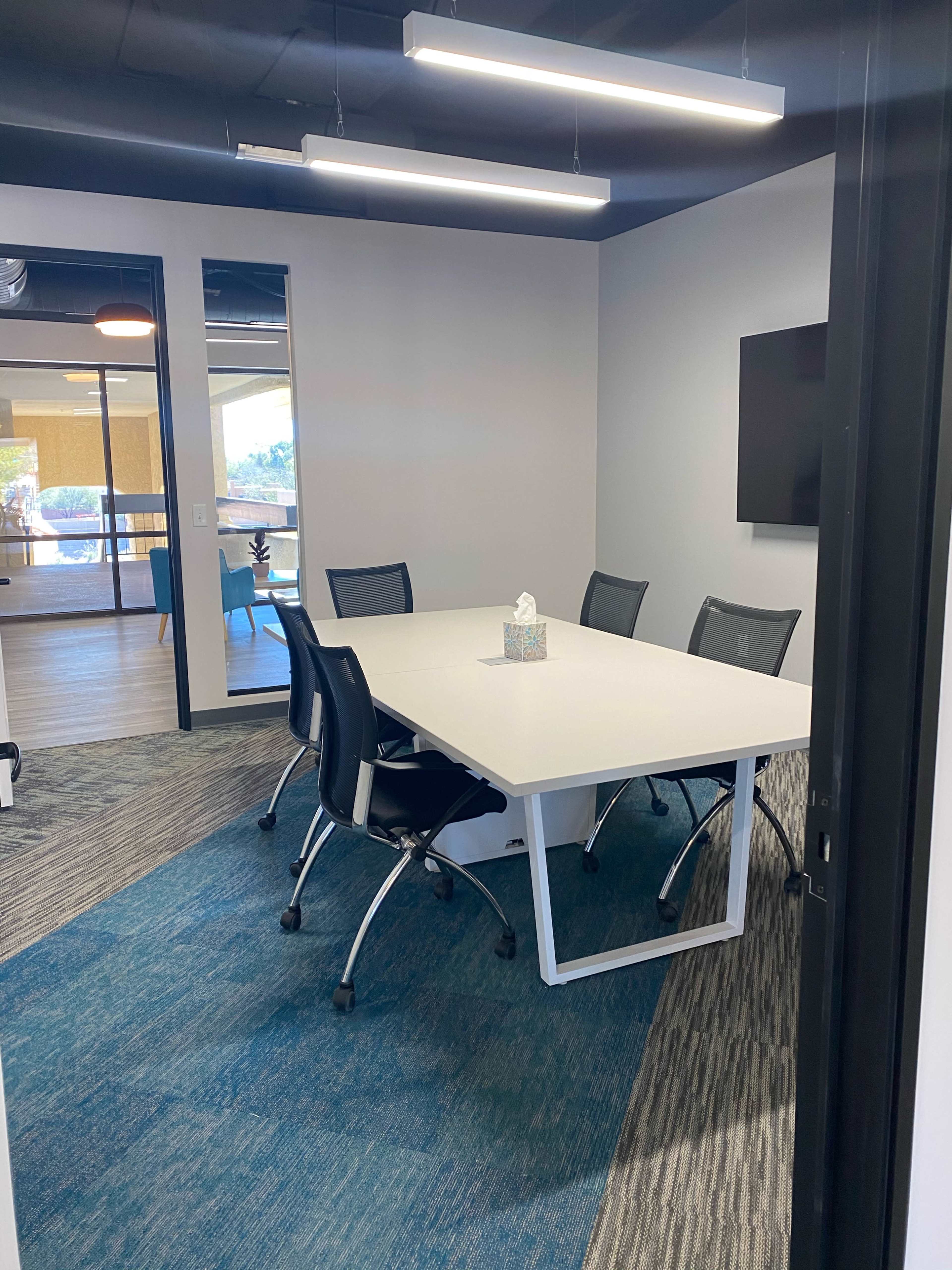 A modern conference room features a large white table surrounded by six black chairs, with a tissue box in the center and a wall-mounted screen.