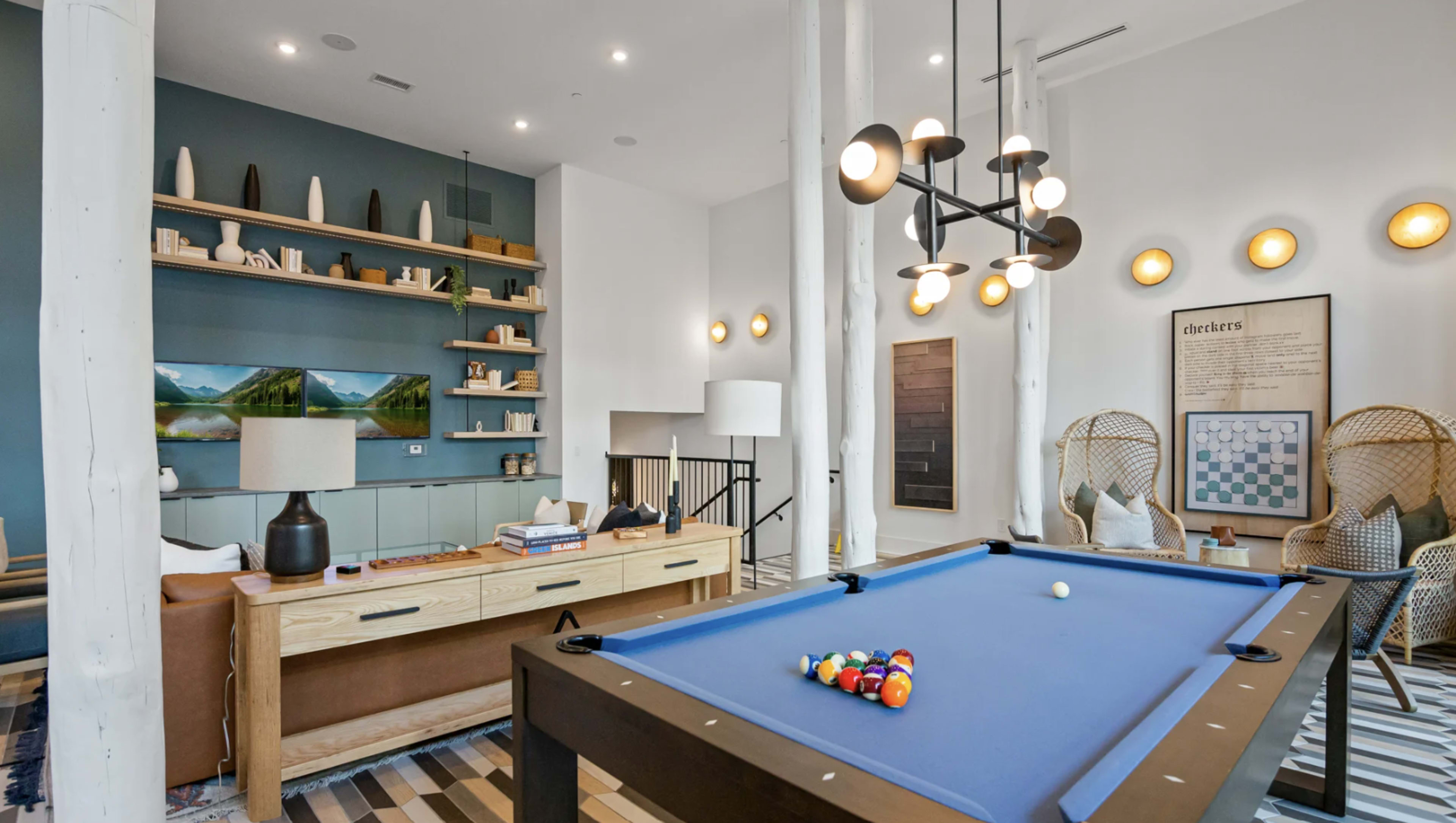 The image shows a modern game room featuring a blue pool table, a wooden console table, decorative shelving, and stylish lighting fixtures.