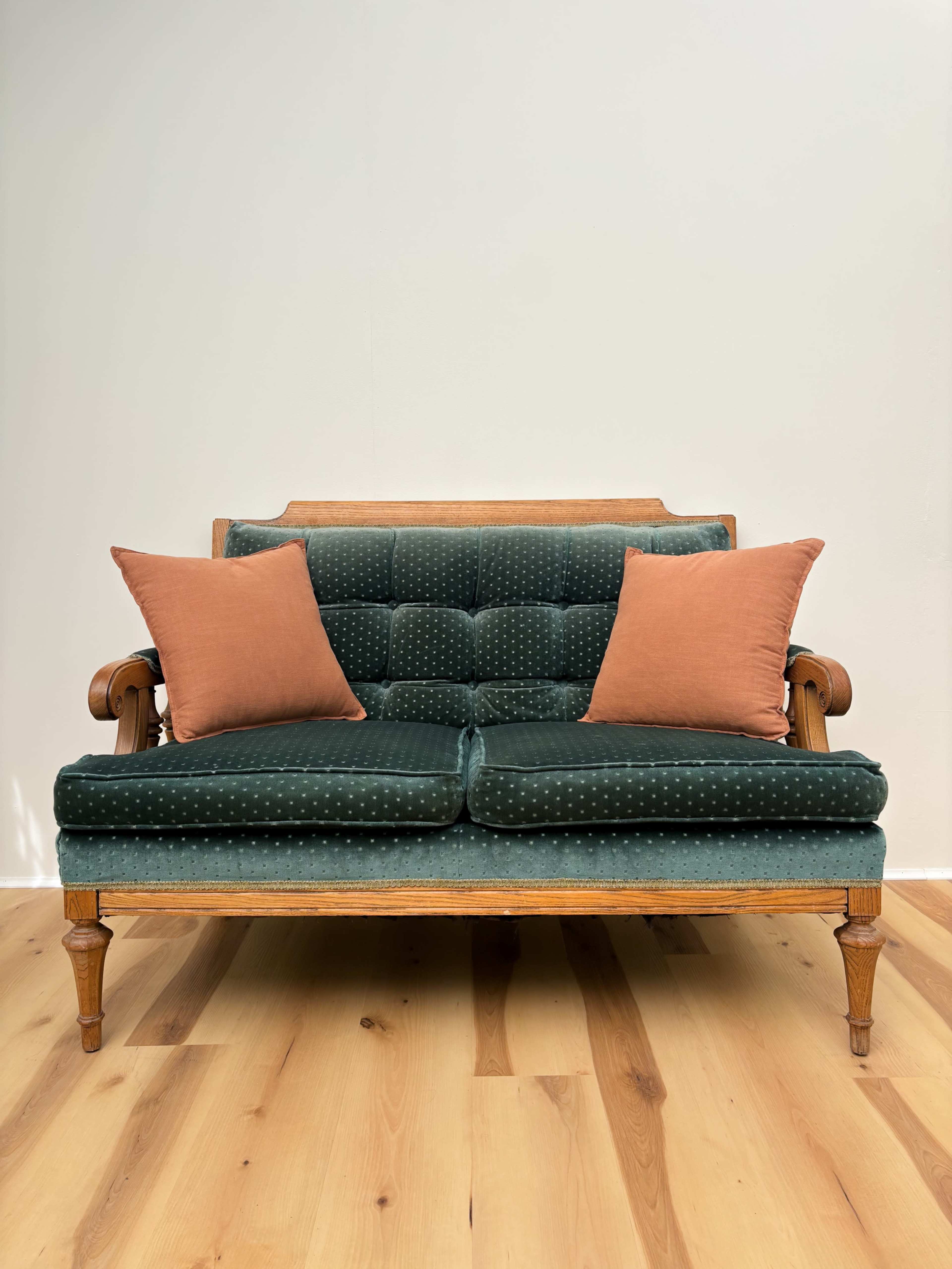 The image features a vintage-style green velvet sofa with two orange cushions, set against a plain white wall and a wooden floor.