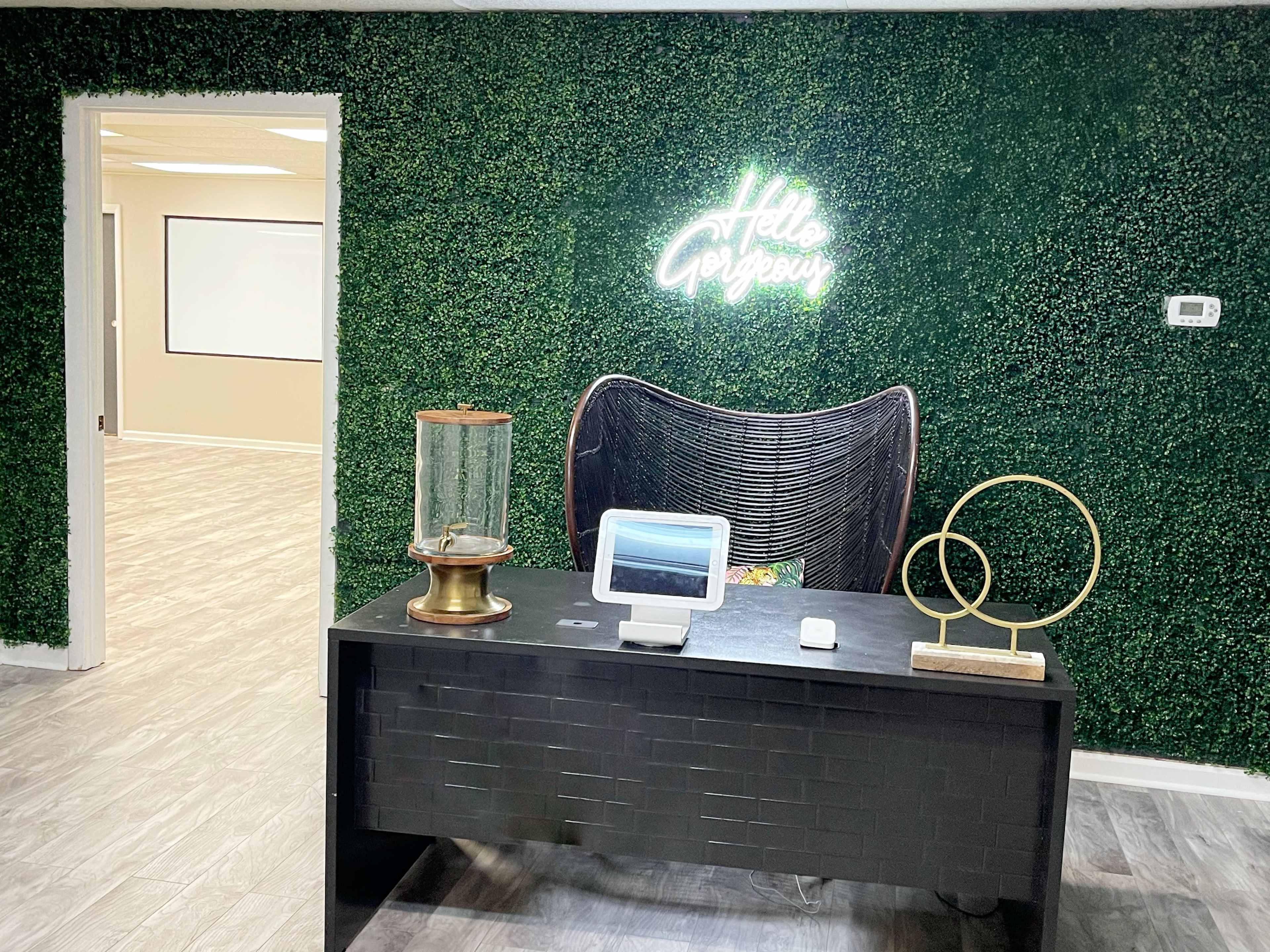 A modern office reception area with a black desk, decorative items, and a green moss wall featuring a neon sign that reads "Hello".