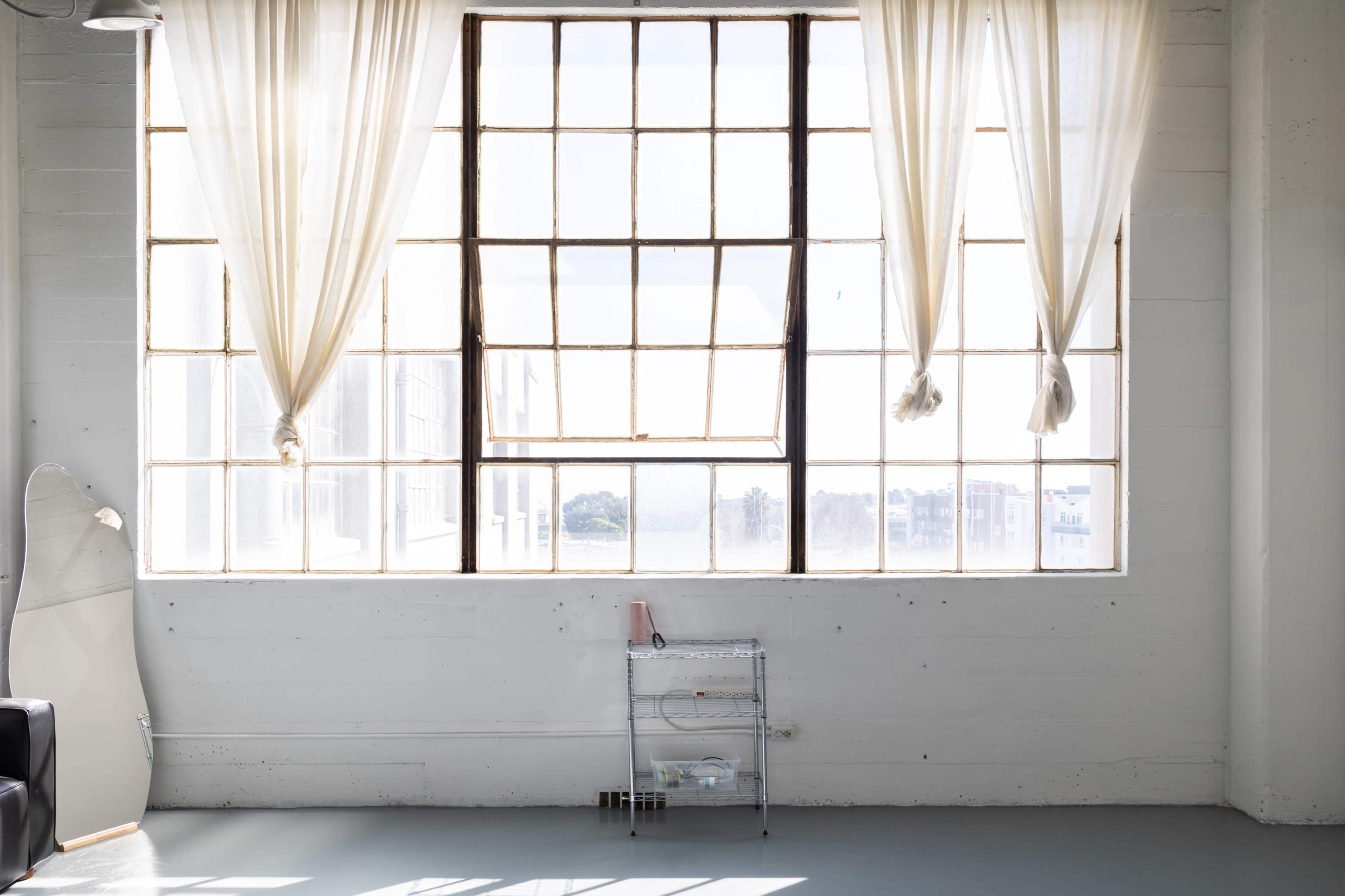 A large window with sheer curtains overlooks a minimalist room with light-colored walls and a small metal cart.