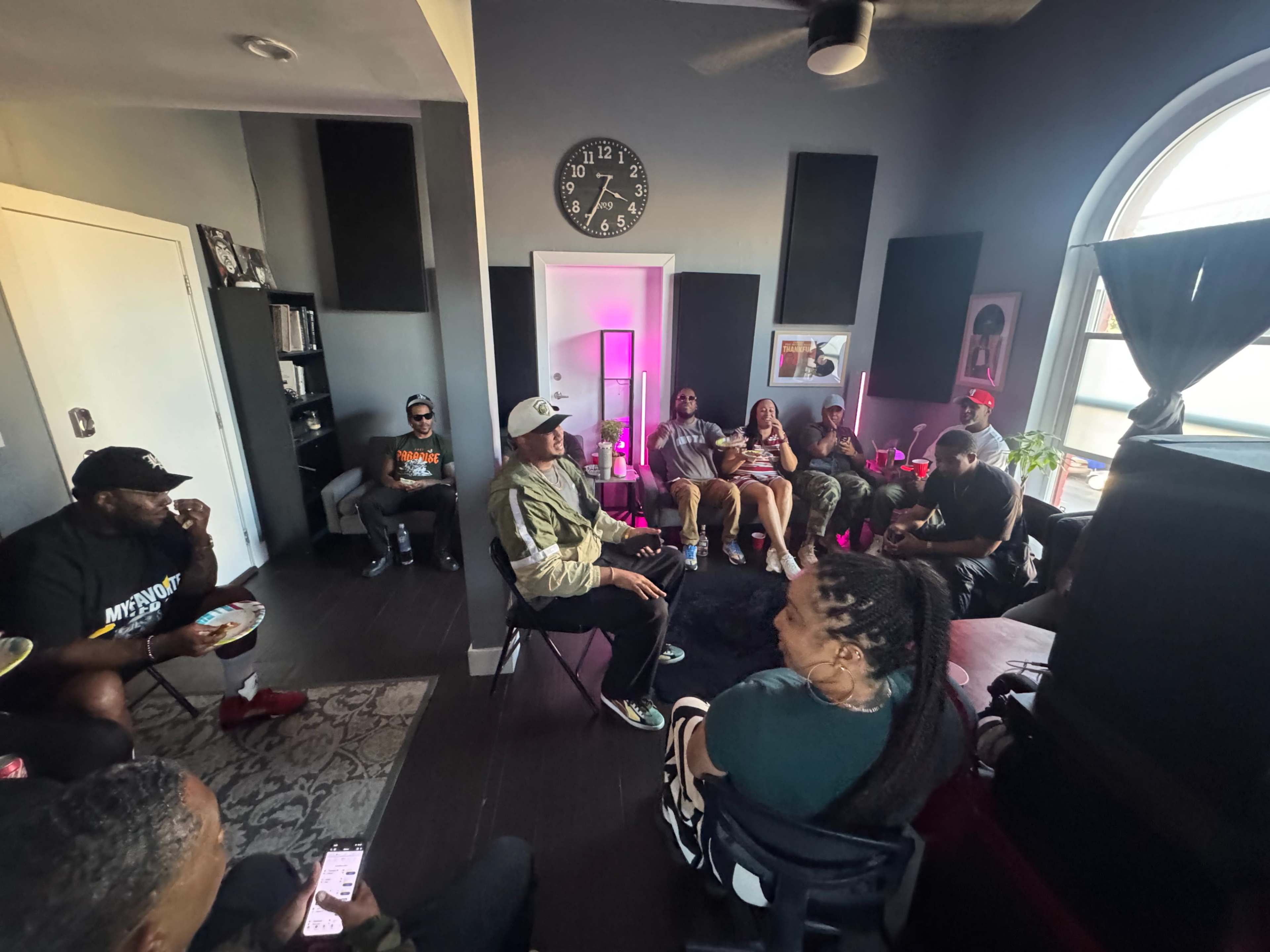 A group of people are seated in a modern living room, engaging in conversation and activities while snacks are visible on a table.