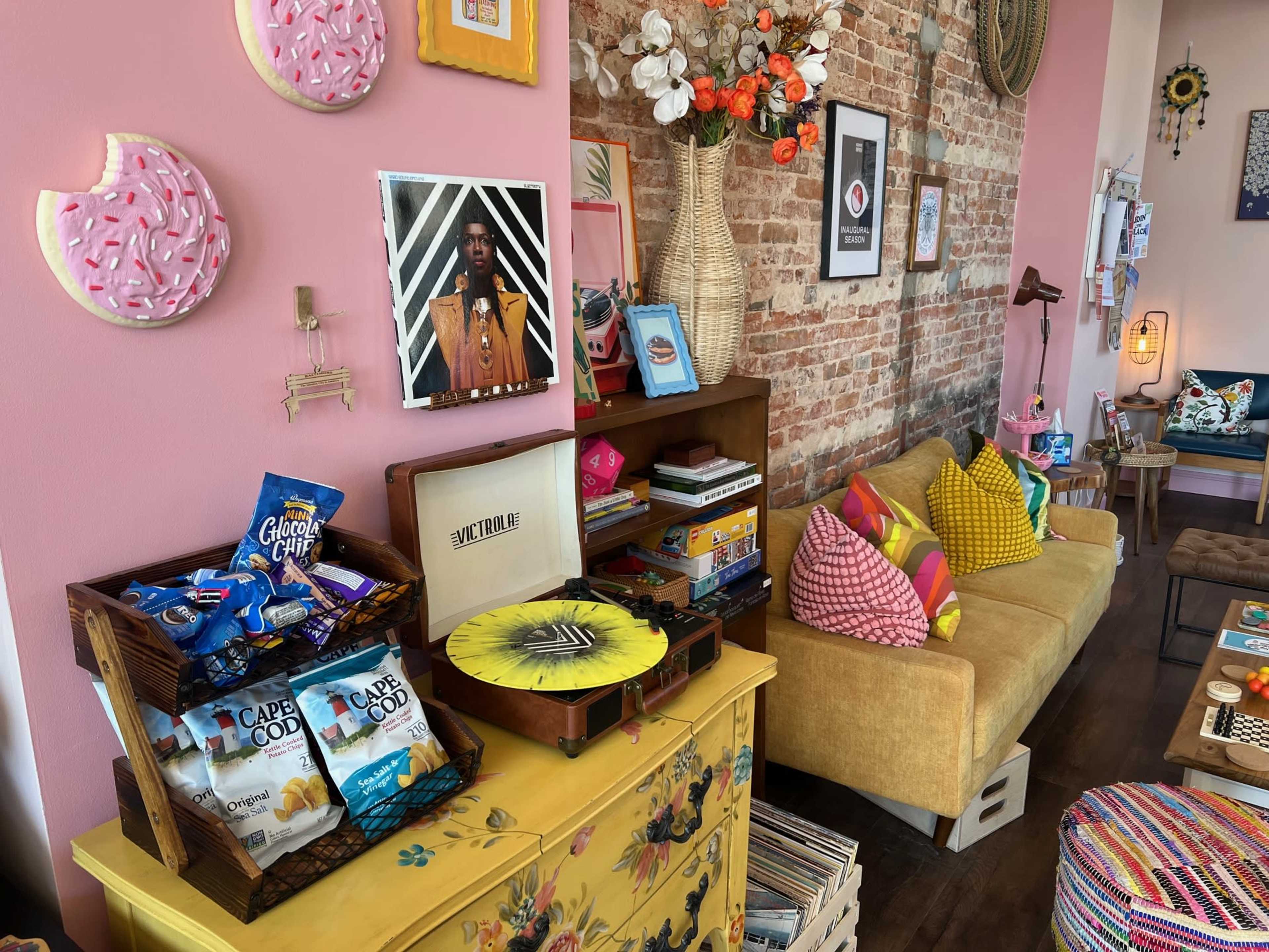 The image features a colorful interior of a cozy café or living space, with a vintage record player, snacks on display, and bright cushions adorning the sofa against a brick wall.