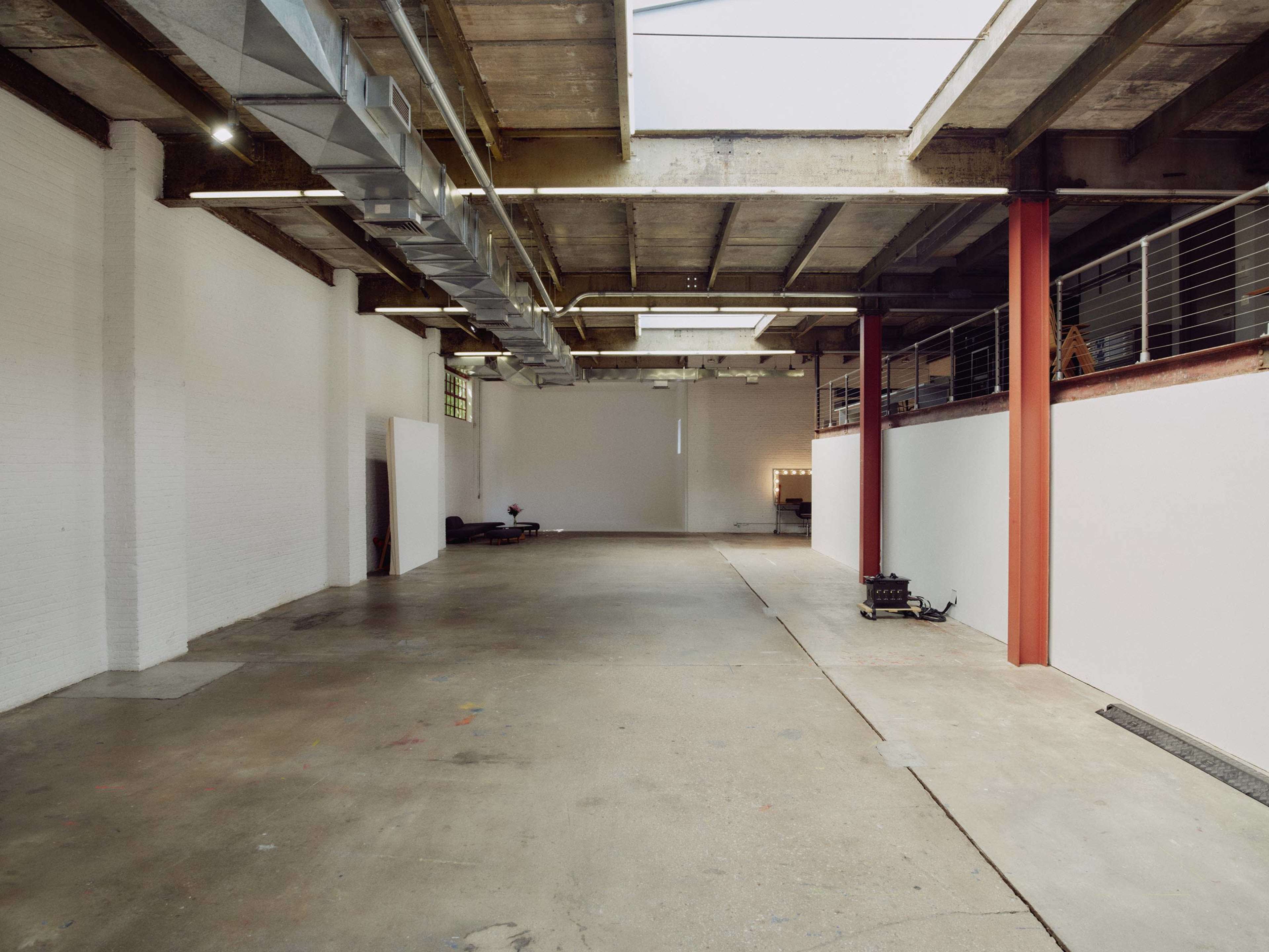The image shows a spacious, empty industrial-style room with concrete flooring, exposed beams, and a large window allowing natural light.