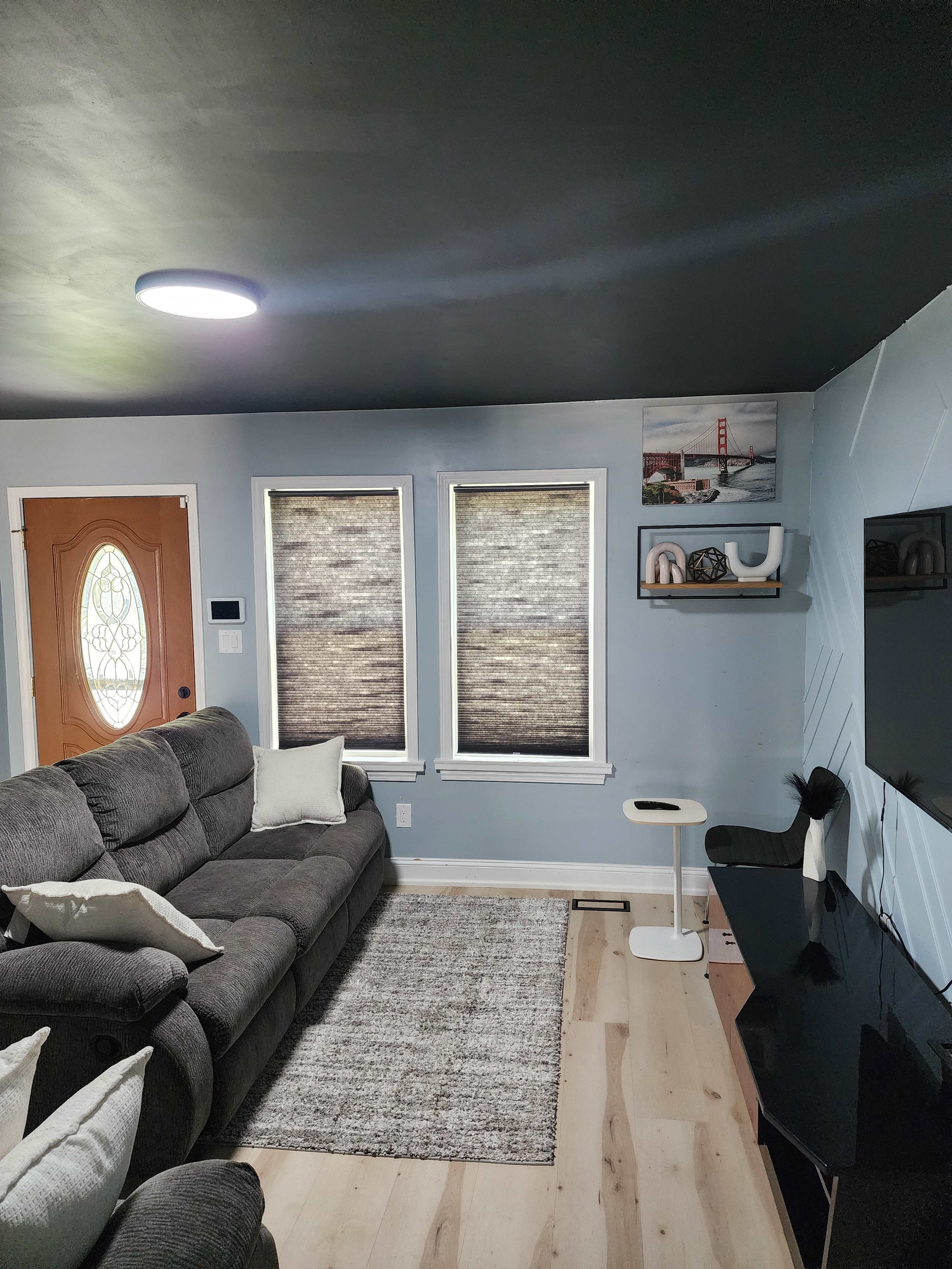 The image shows a living room with a gray sectional sofa, a light-colored rug, and a wall-mounted television beneath a shelf displaying decorative items.