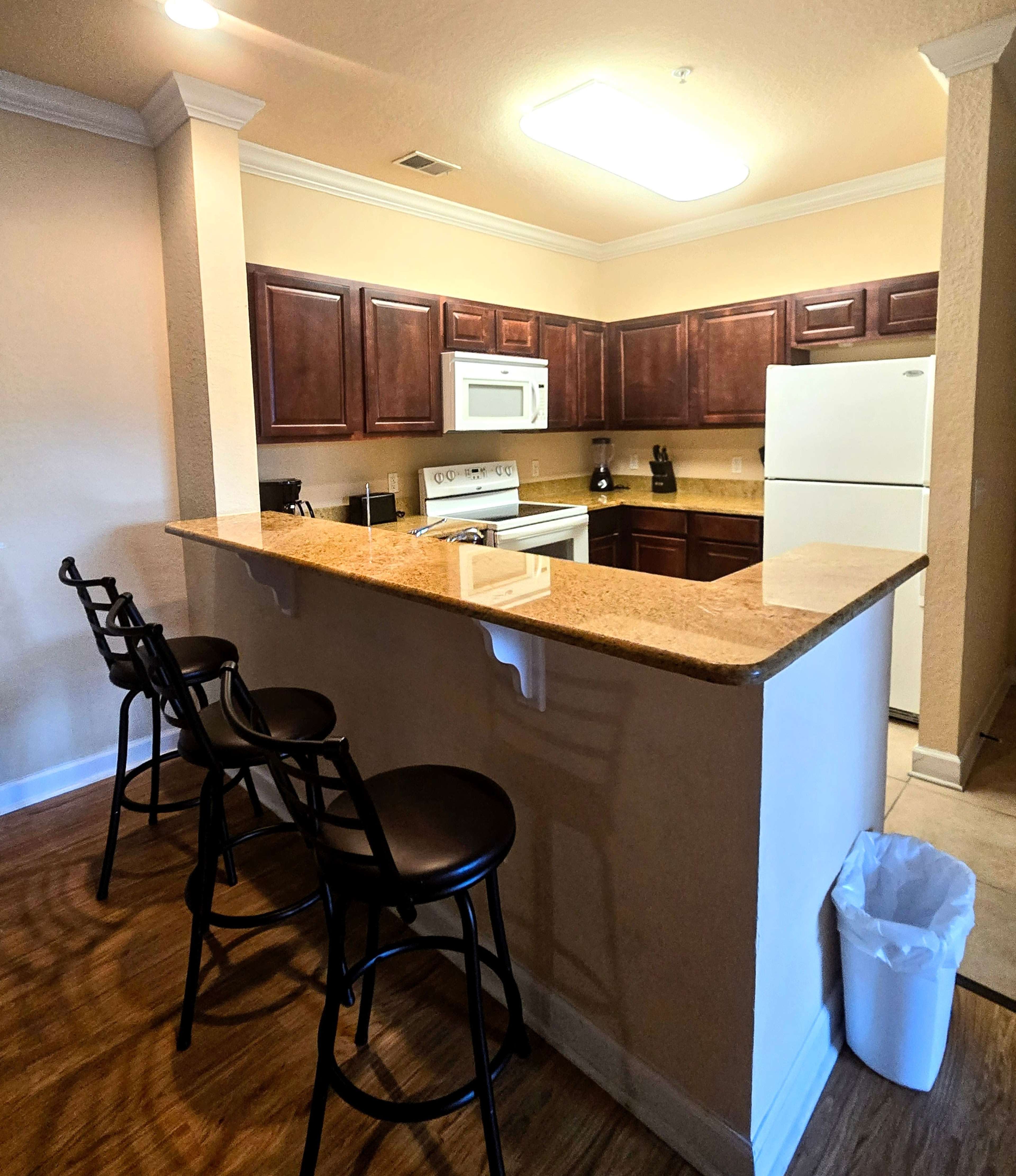 A modern kitchen features dark wooden cabinets, a granite countertop, and appliances including a microwave, stove, and refrigerator, with four black barstools at the breakfast counter.