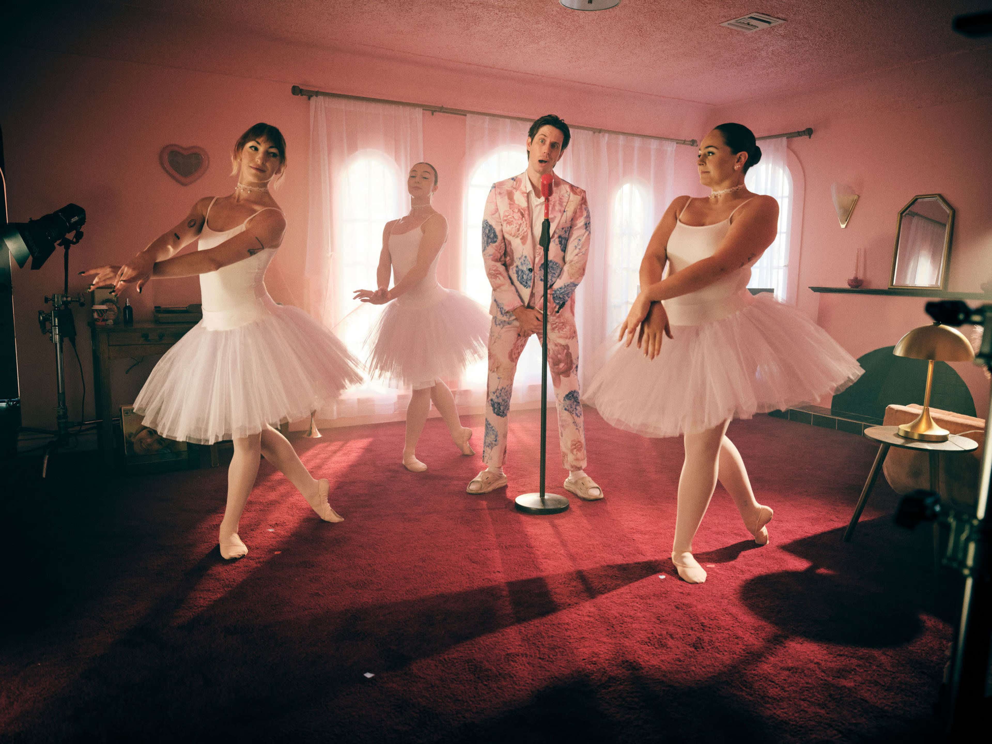 A man in a patterned suit stands at a microphone, surrounded by three ballet dancers in tutus, all set in a pink room with soft lighting.