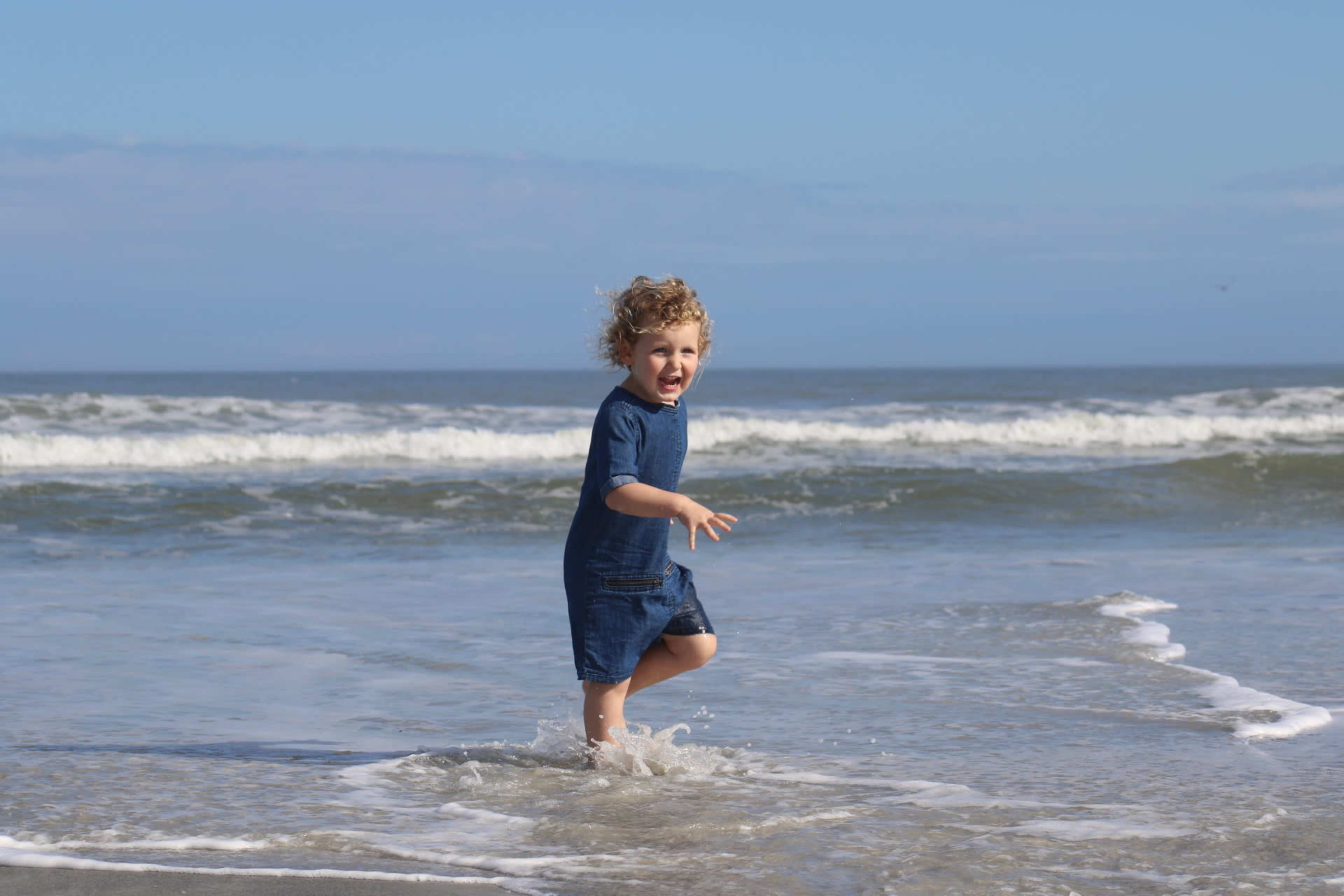 A child with curly hair joyfully splashes in the shallow waves at the beach.