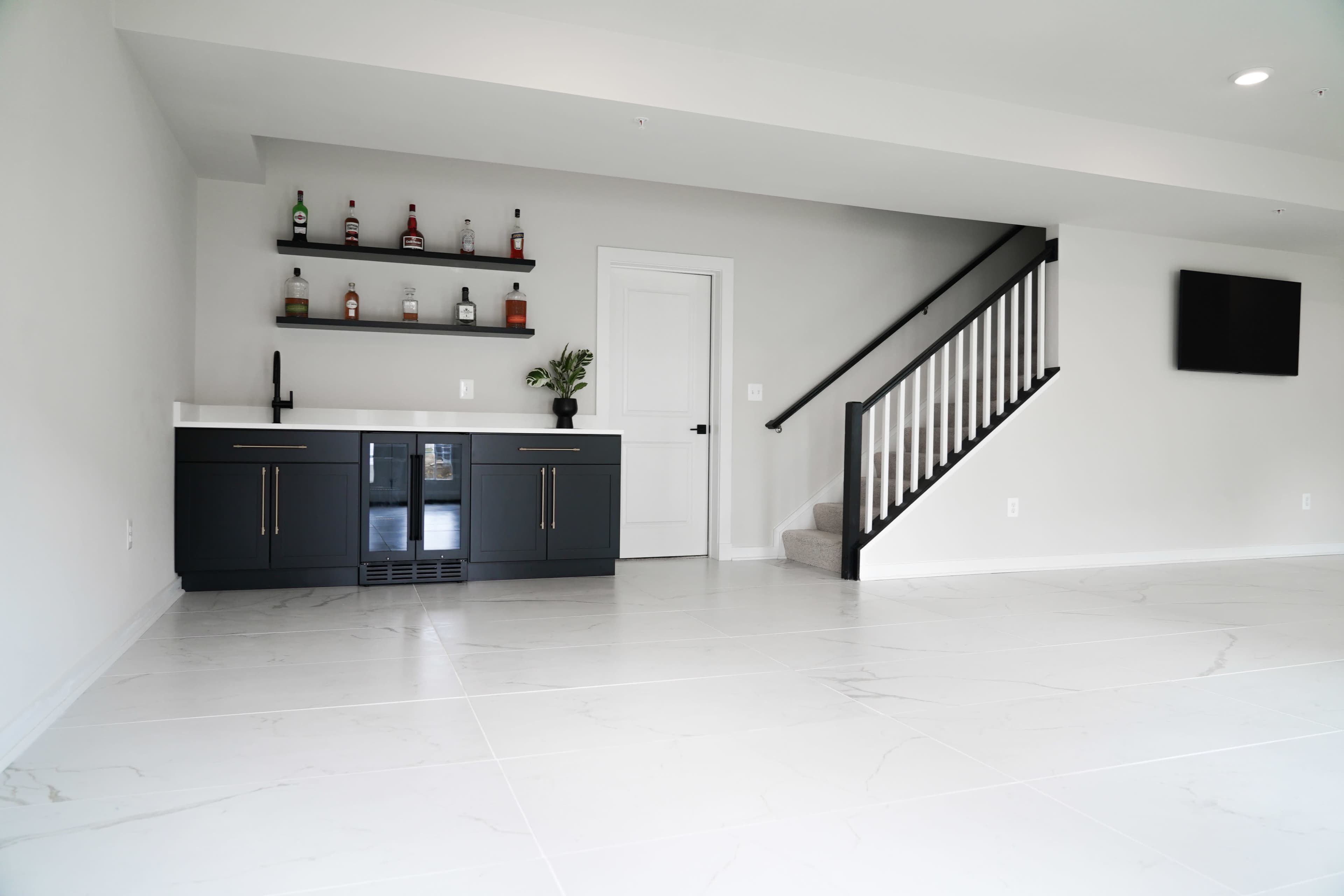 The image shows a modern interior space featuring a sleek bar area with a beverage cooler and shelves, alongside a staircase leading to an upper level.