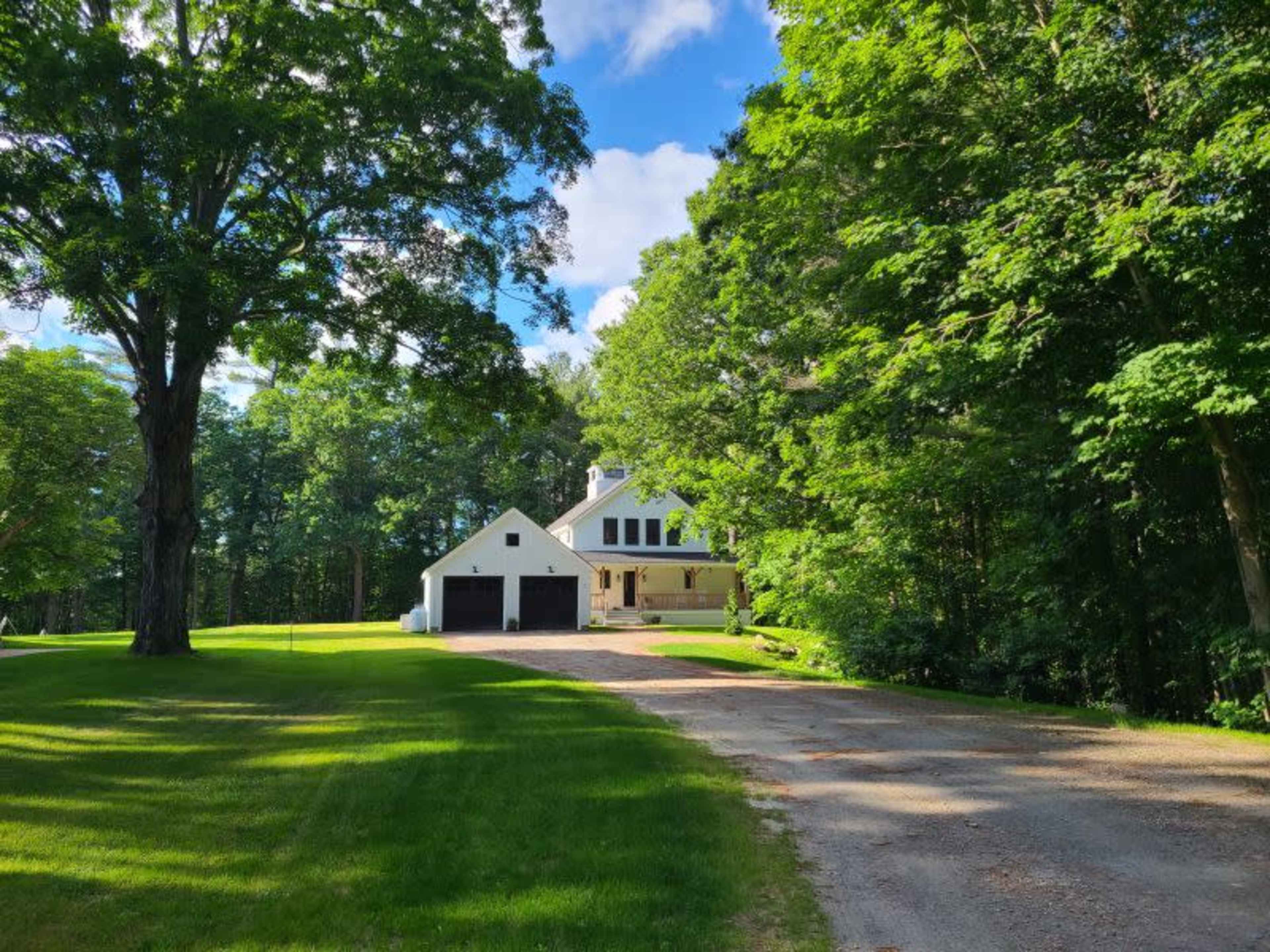 A white house with a black garage is situated along a gravel driveway surrounded by lush green trees.