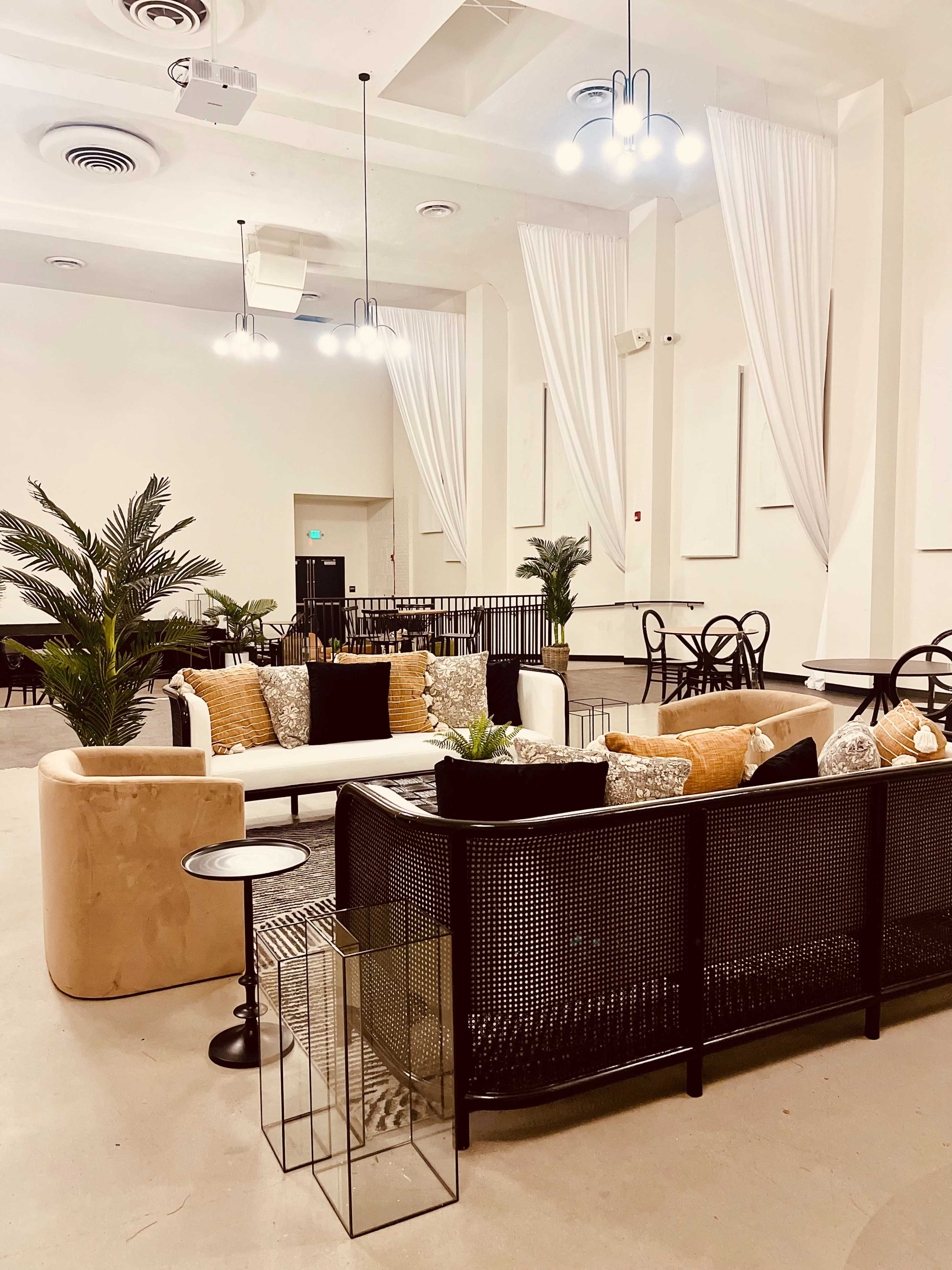 A modern, spacious lounge area furnished with sofas, potted plants, and tables, illuminated by overhead lights.