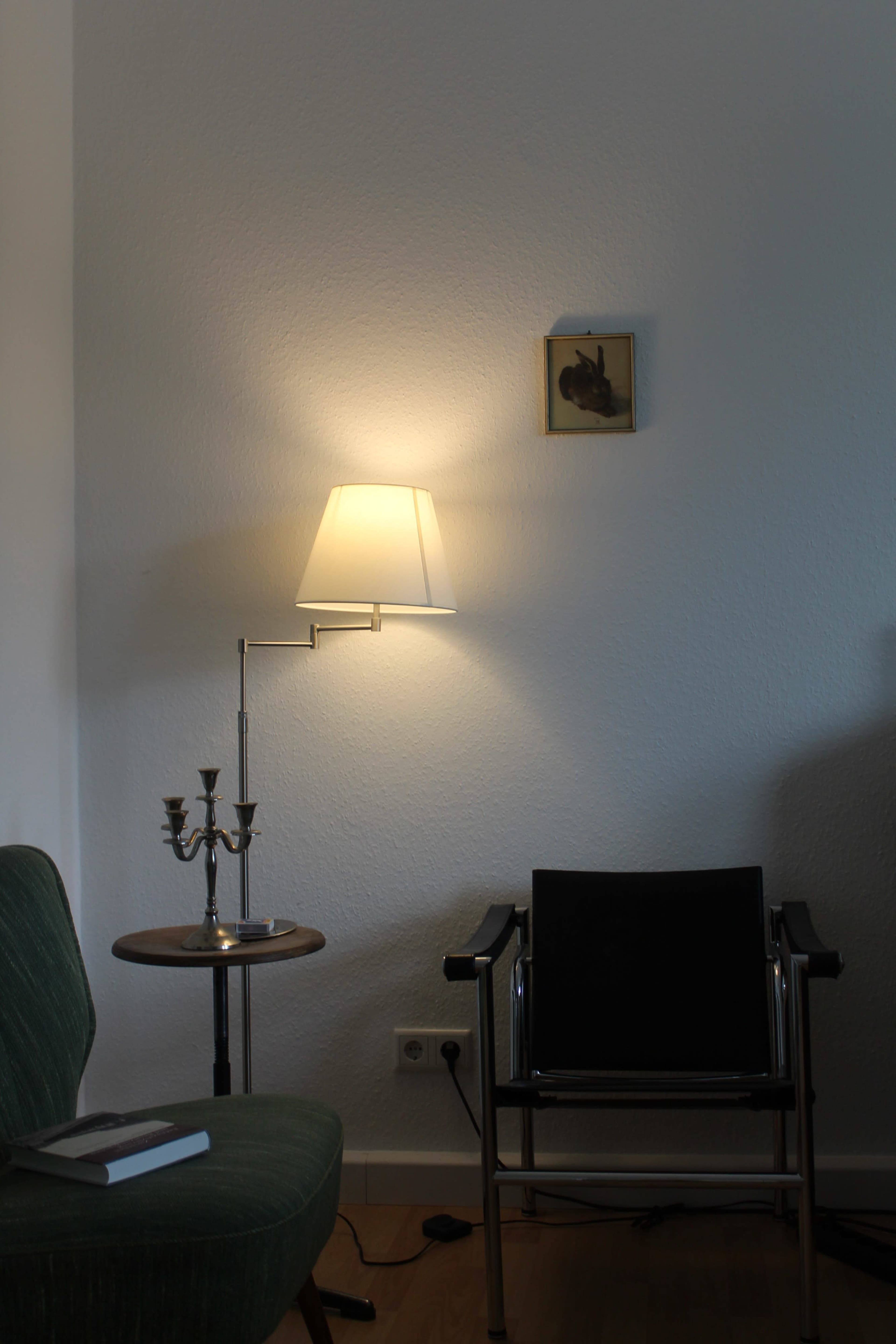 A green upholstered chair and a black metal chair sit beside a small table with a candlestick, illuminated by a lamp on the wall, with a framed picture hanging above.